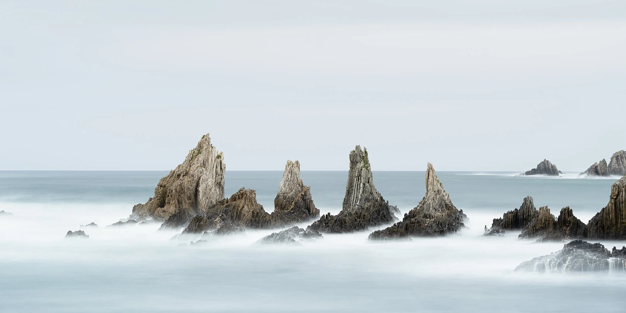 Misty seascape with jagged sea stacks rising from calm ocean, long-exposure coastal rocks under soft sky