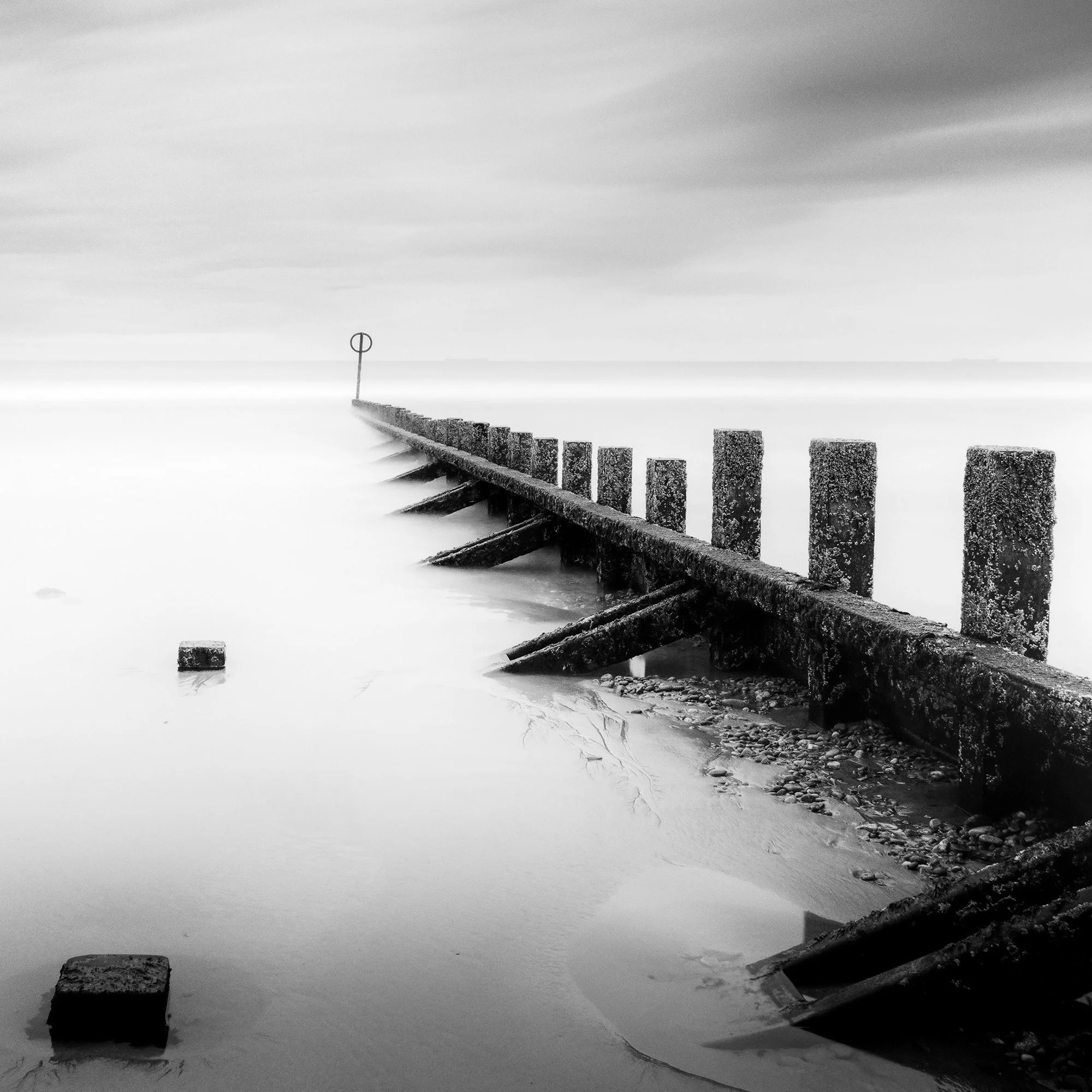 © 2012 Gerald Berghammer - Black and white landscape photography. A wooden wave breaker extending into a calm body of water, with a circular marker at the end. Print detail 2