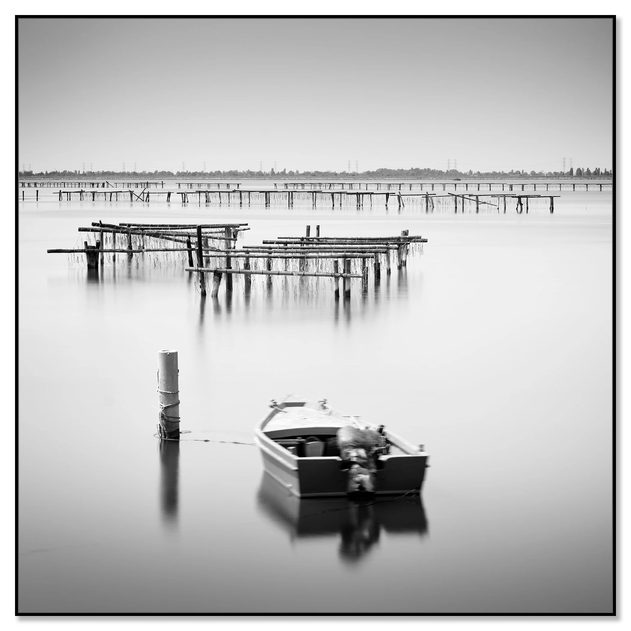 Black-and-white seascape photograph of a small boat tied to a post on calm water, with wooden aquaculture piers in Italy – framed ArtBox black