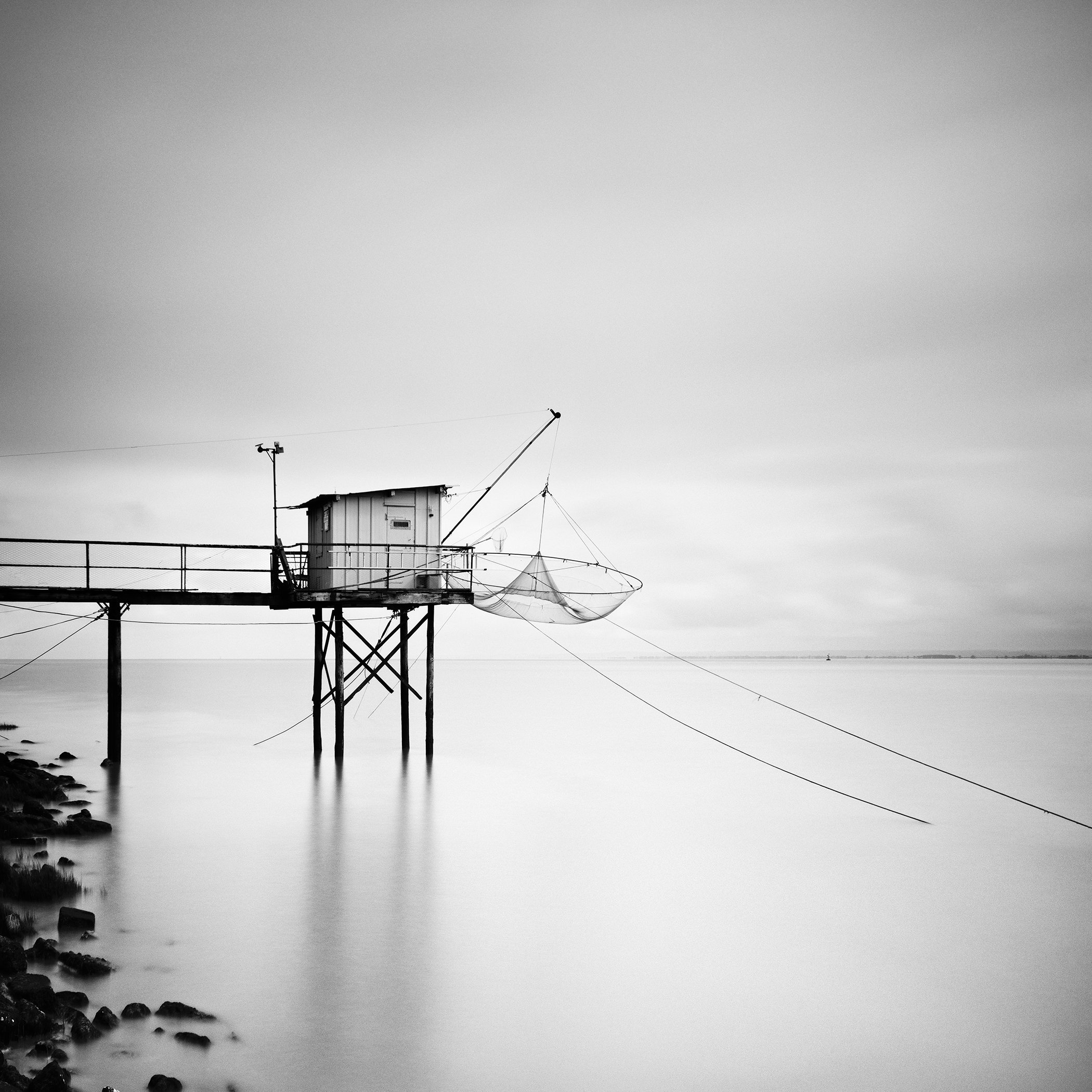 Minimalist black-and-white long-exposure of a stilted fishing hut with nets over a glassy calm sea, rocky shoreline in the foreground