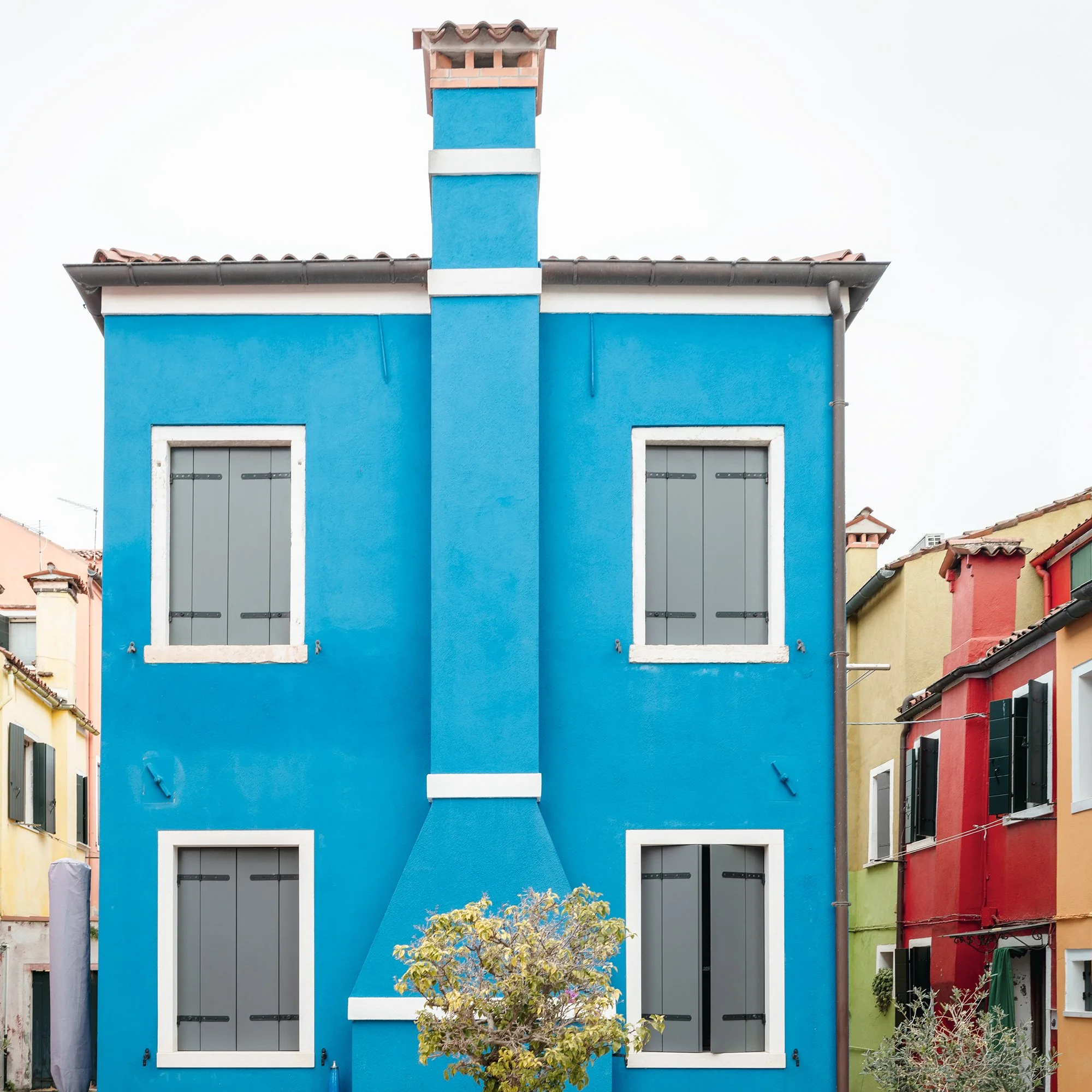 © 2025 Gerald Berghammer - Color cityscape photo. Colorful row houses with a prominent blue house in the center, potted small trees in front, and a paved courtyard. Print detail 2
