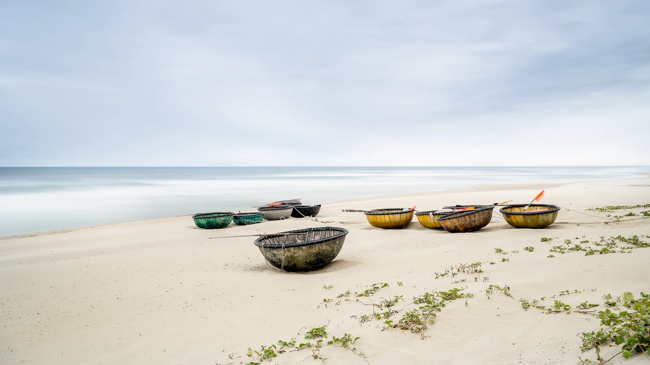Calm sandy beach with colourful fishing boats on the shore, a smooth sea and pale sky on the horizon