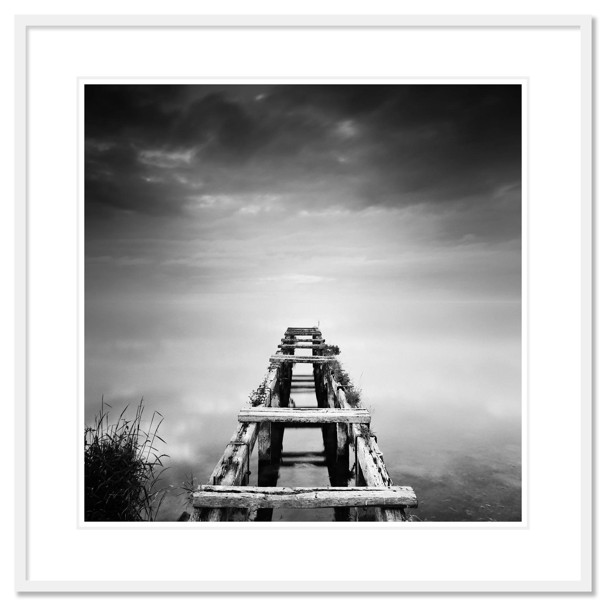 Gerald Berghammer - Black and white waterscape photography. An old abandoned wooden dock extending into a calm body of water under a cloudy sky. Classic framed white