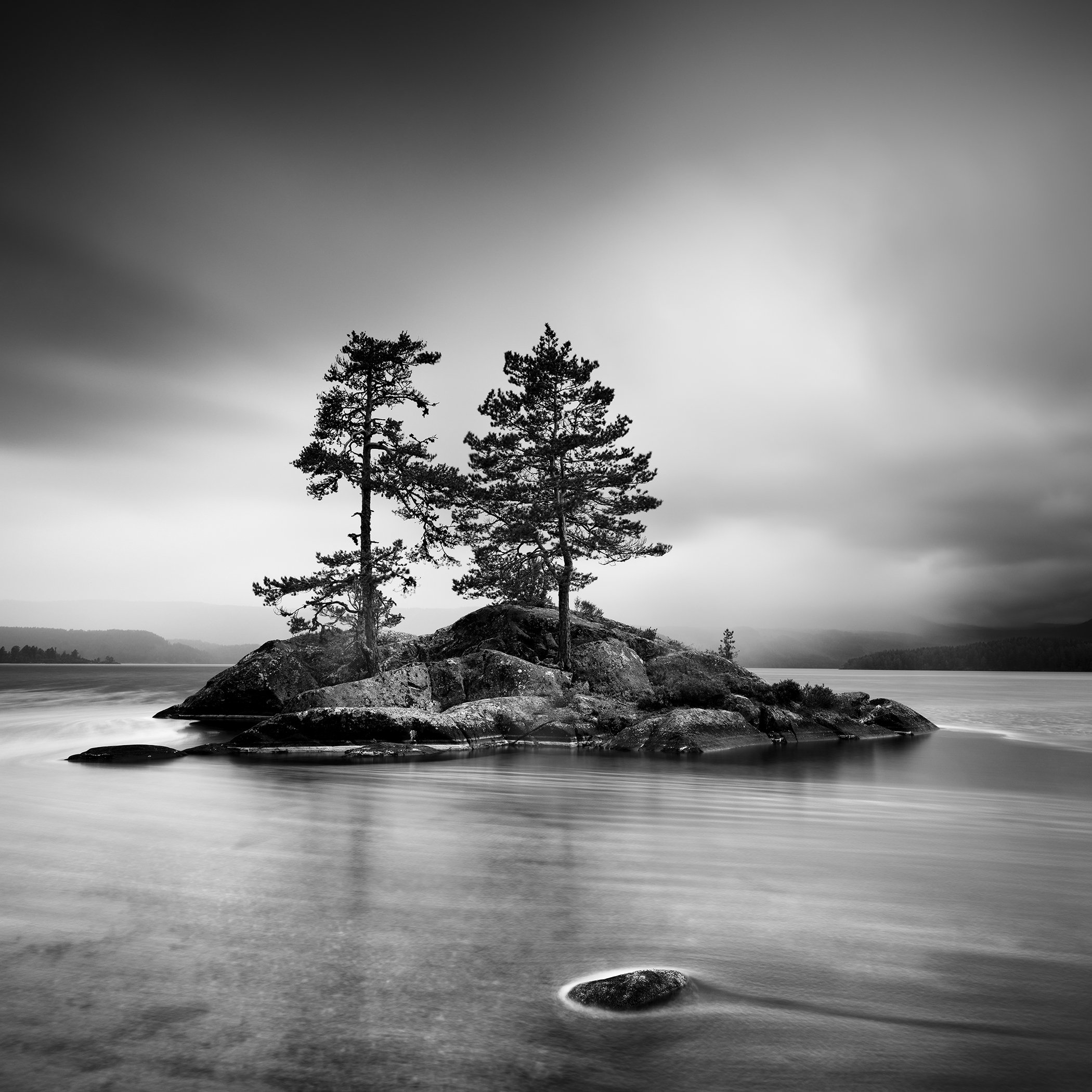 Monochrome long-exposure photo of two trees on a small rocky islet surrounded by still water and misty hills