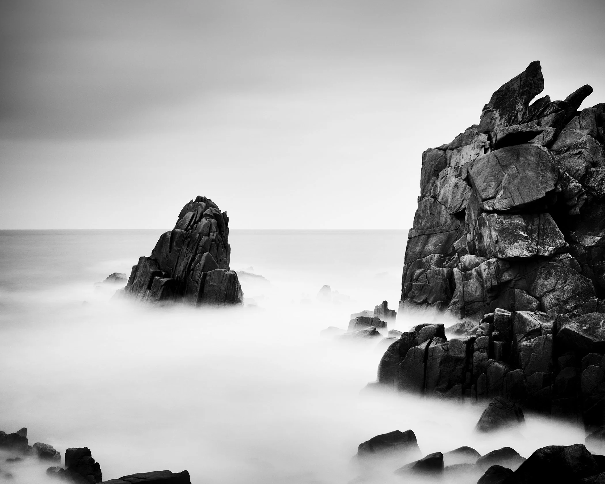 Monochrome coastal landscape featuring dramatic rock formations and smooth fog-like water