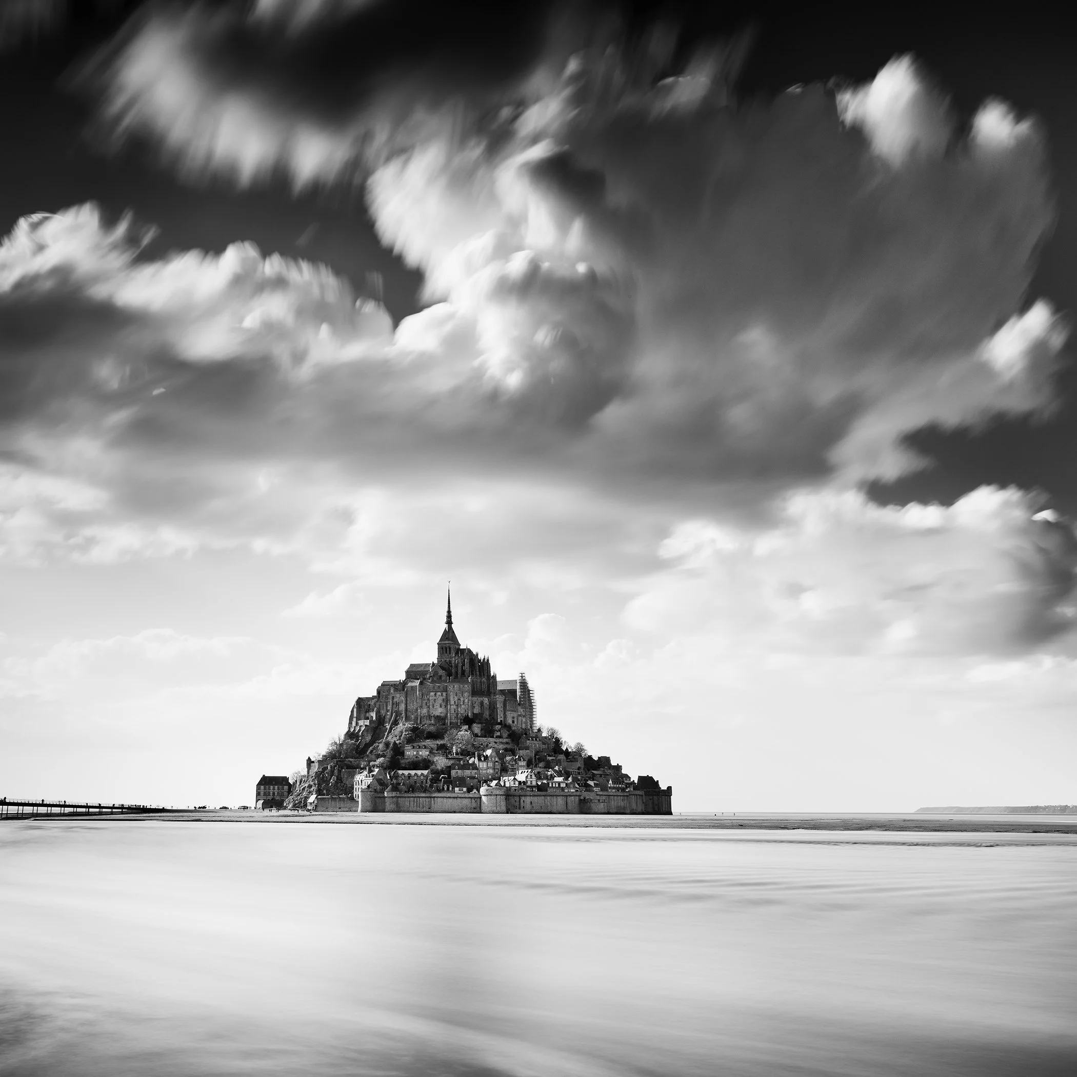 Mont Saint-Michel in France captured in black and white, with sweeping clouds and smooth sea in the foreground.