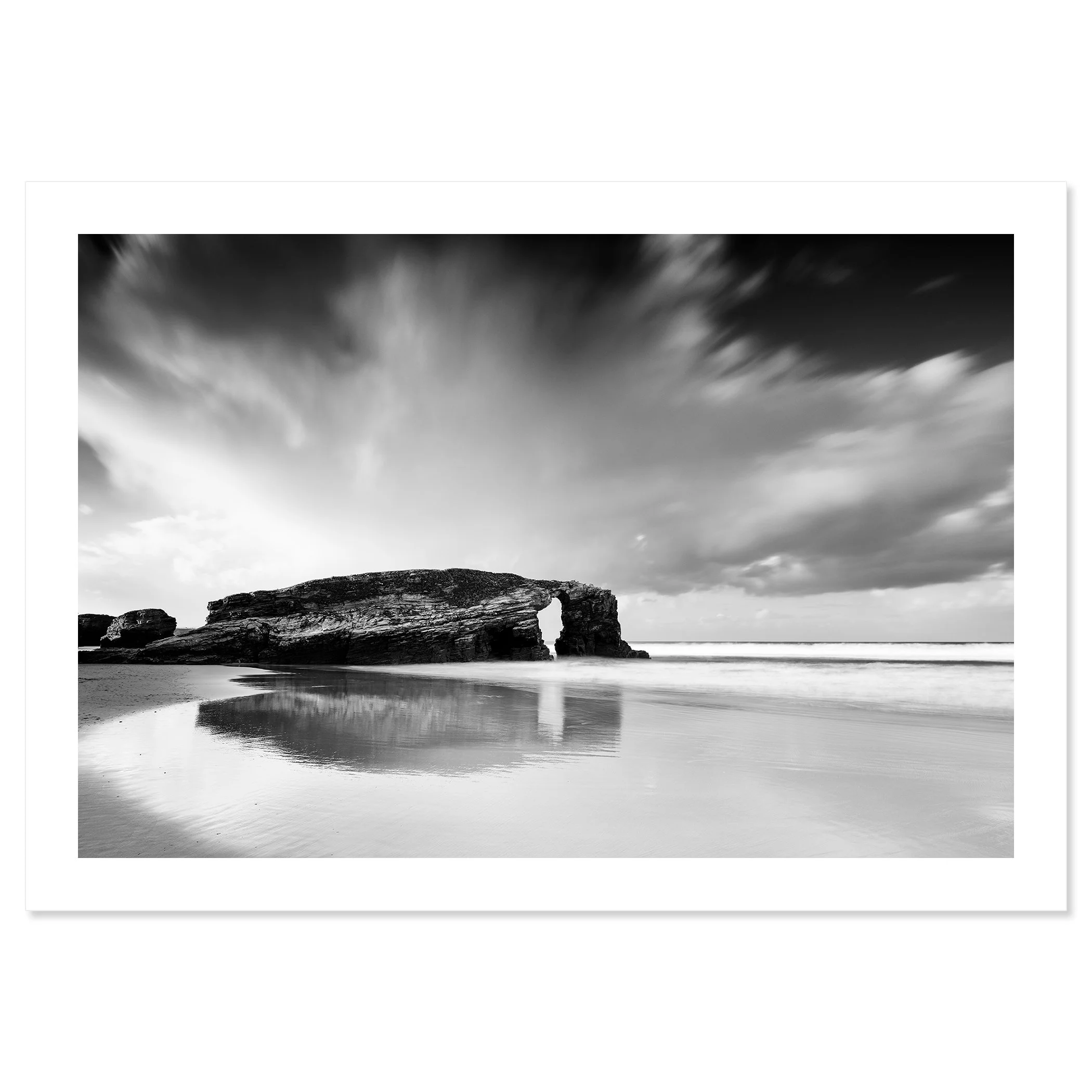 Gerald Berghammer - Black and white Photography. A rocky beach with a natural arch formation, reflecting in the wet sand, under a sky with moving clouds. Fine art print only