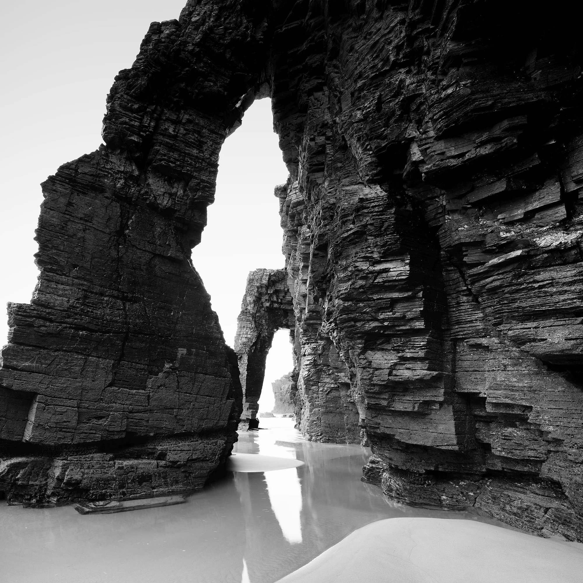 Gerald Berghammer - Black and white beach seascape photography. Large natural rock arch formations on a beach with water reflecting the rocks. Print detail 2