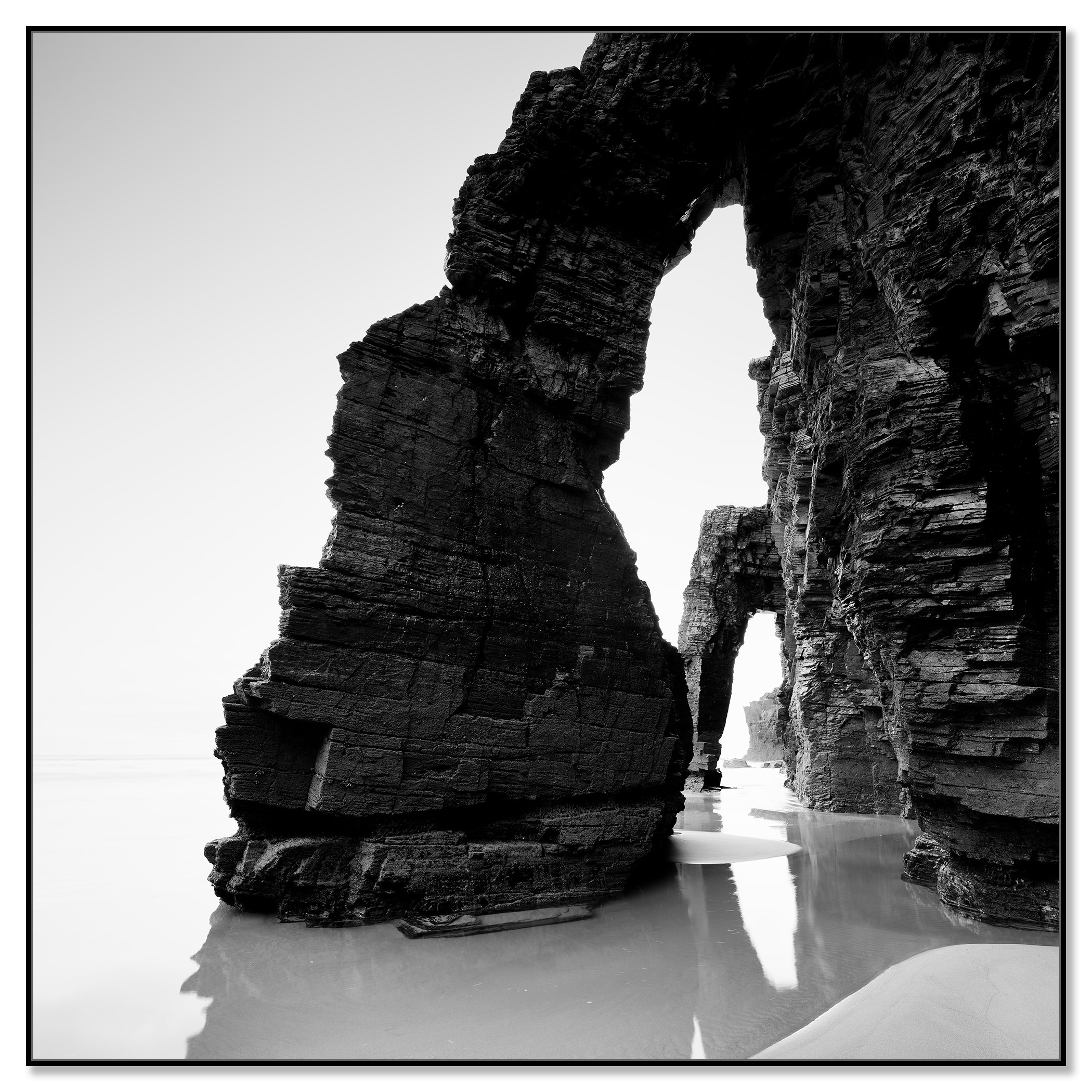 Black and white photo of dramatic sea arches and layered rock cliffs on a quiet sandy beach – framed ArtBox black