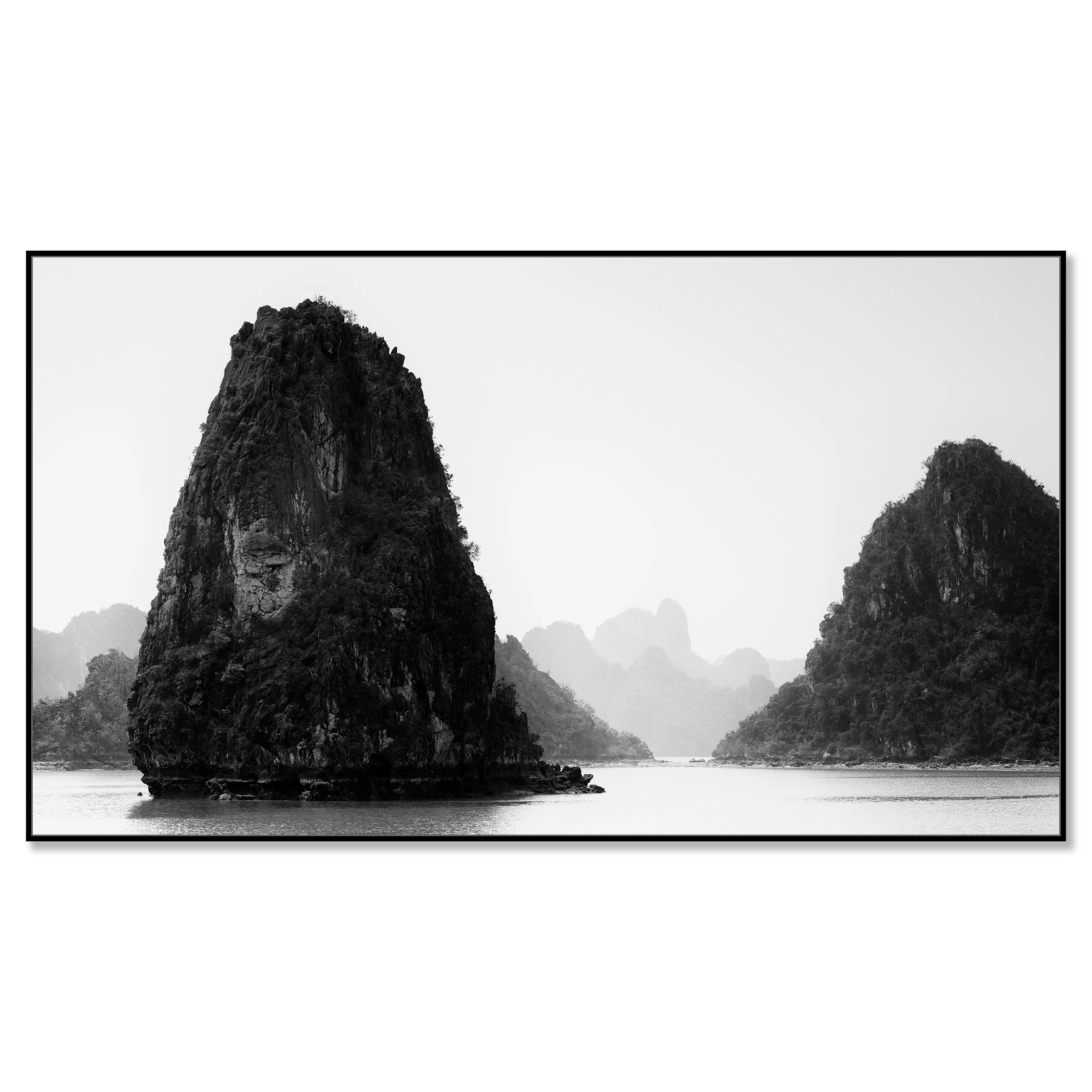 Black-and-white seascape in Vietnam showing limestone karst formations rising from calm water, with misty mountains in the background – framed ArtBox black