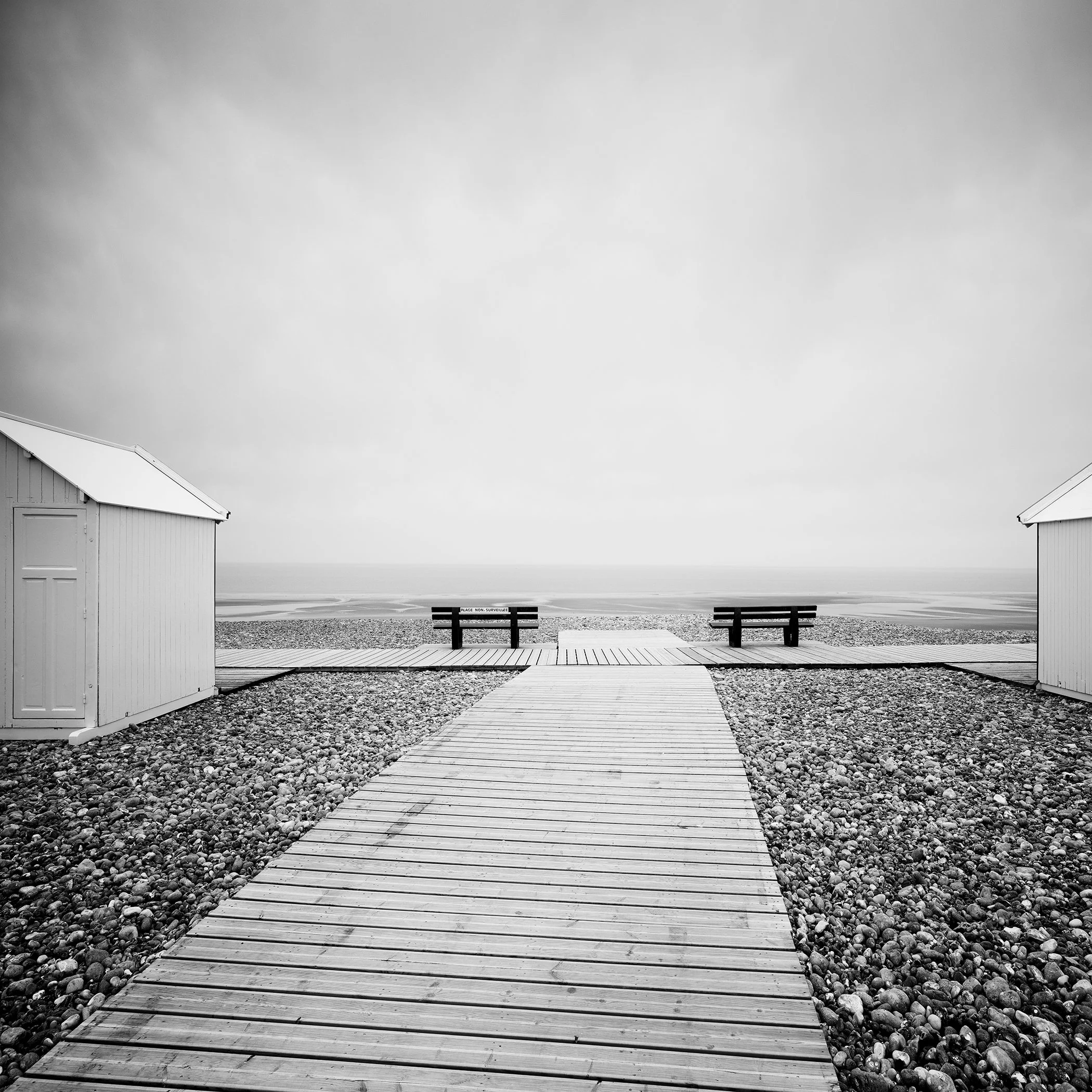 Monochrome coastal scene with a timber walkway, pebble shore, beach huts and benches overlooking calm water
