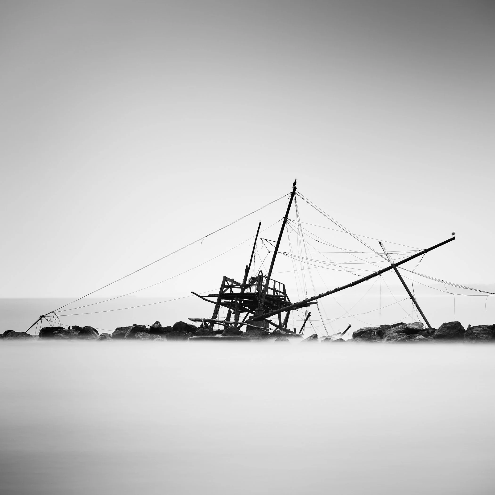 Abandoned fishing hut at Bocca d’Arno in black and white, rising from the calm sea beside a line of rocks.
