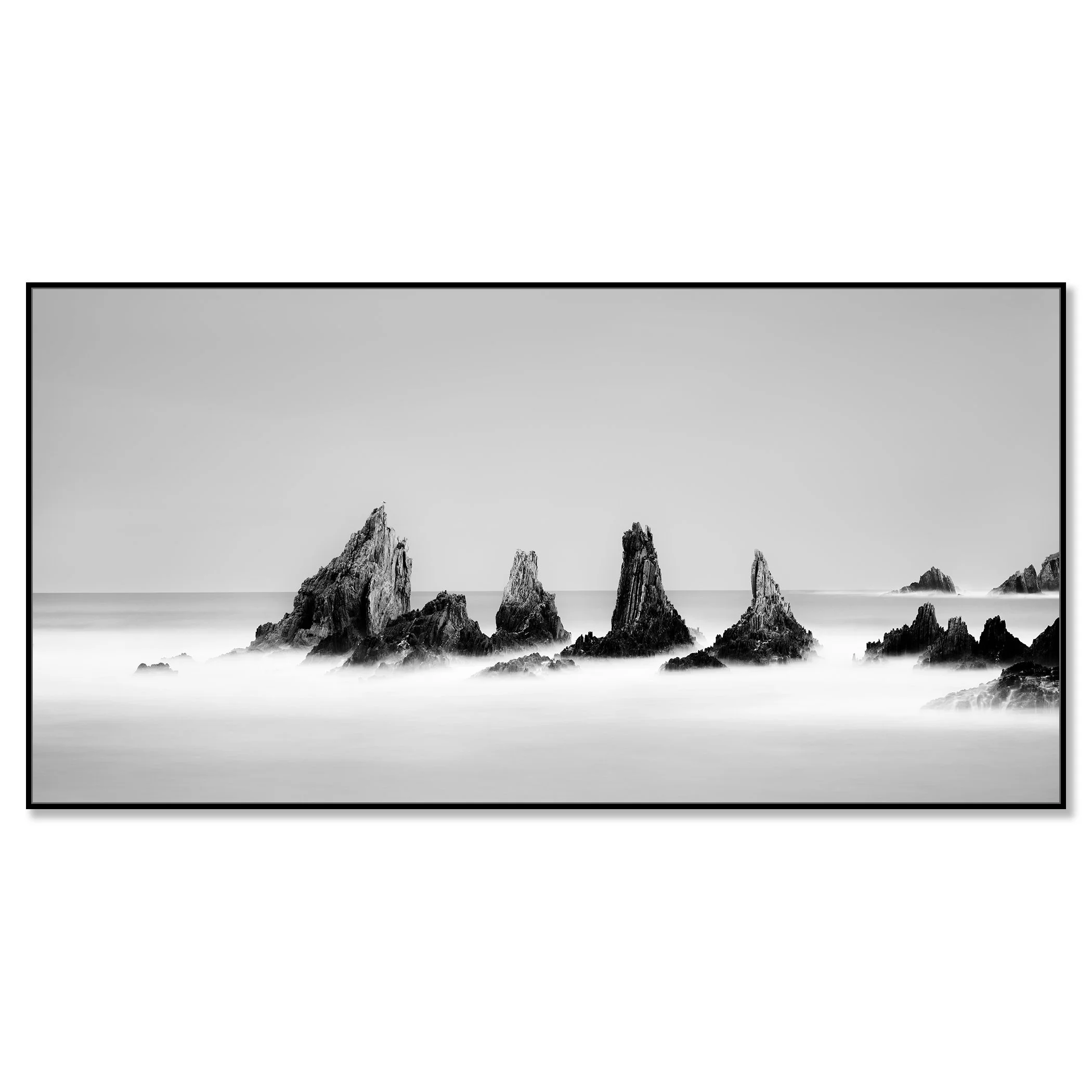 Black-and-white long-exposure photograph of jagged rocks in calm ocean water, misty waves and clear sky – framed ArtBox black