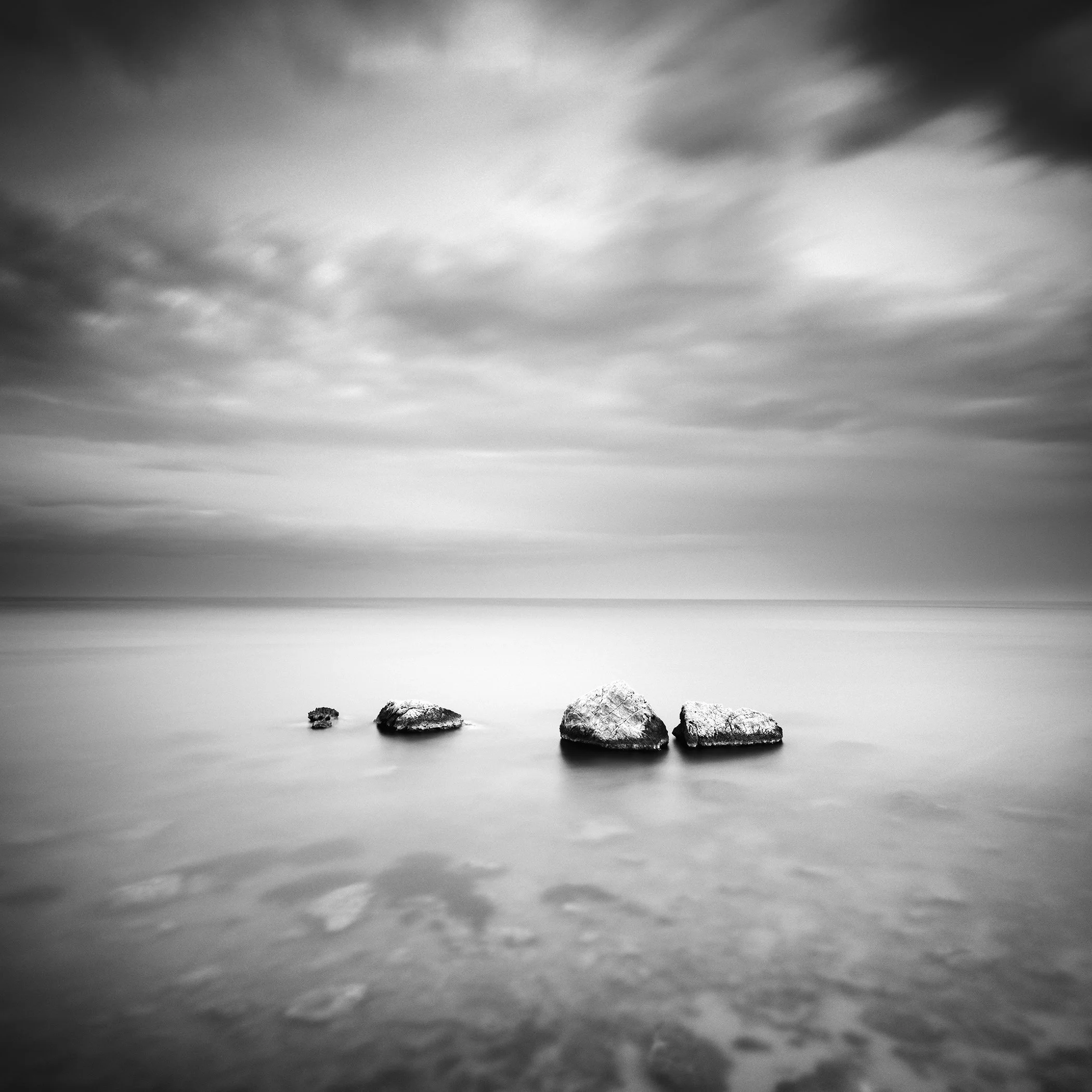 Black and white minimalist seascape with smooth water, stones in the foreground, and a cloudy sky.