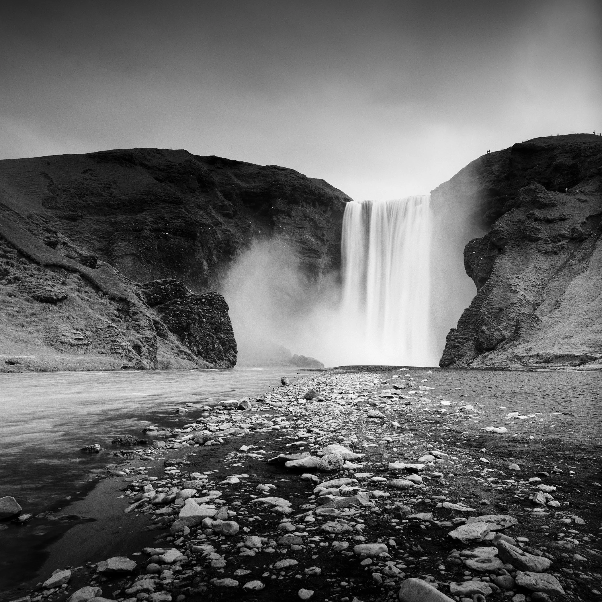 Black and white landscape photo of a powerful waterfall cascading between rocky cliffs with a riverbed in the foreground.