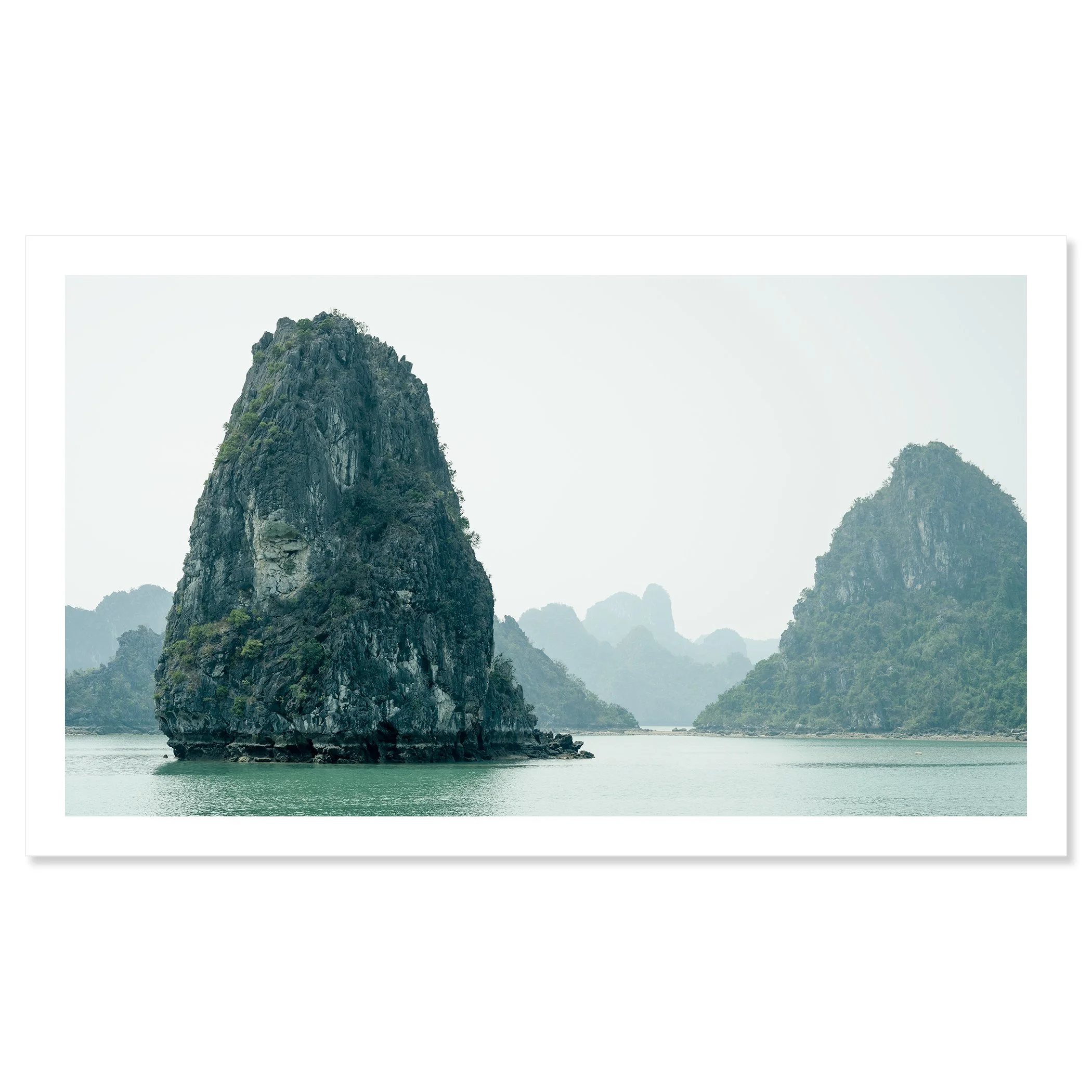 Gerald Berghammer - Color Seascape Photography. Limestone karst formations rise from the water in a tranquil bay, with hazy mountains, Southeast Asia. Fine art print only