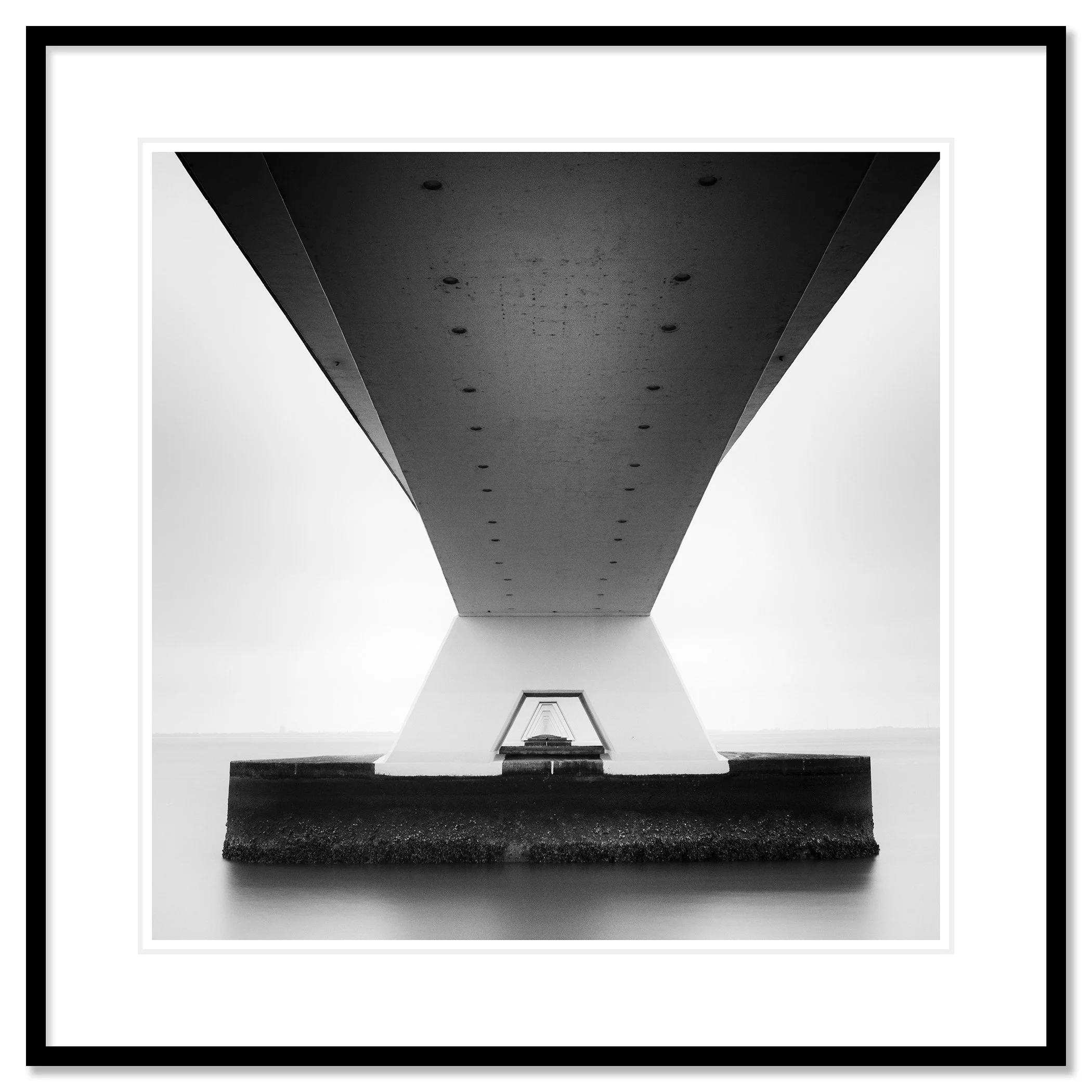 Gerald Berghammer - Black and white cityscape photo. Underneath view of a bridge with large support pillars, showing the underside and the water below. Classic framed black