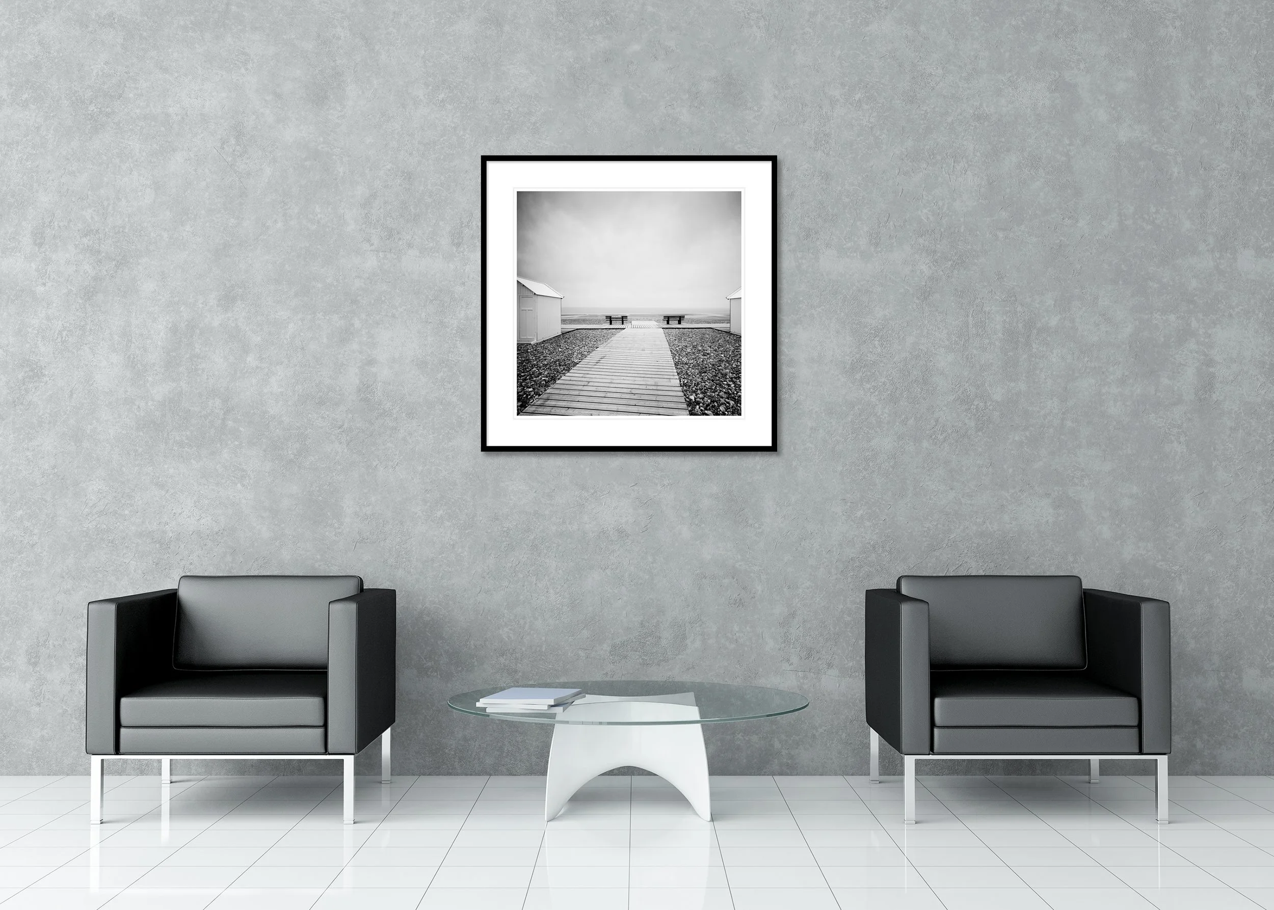 Black and white archival pigment print of a wooden path on a shingle beach between white beach huts, in a classic frame with a mount.