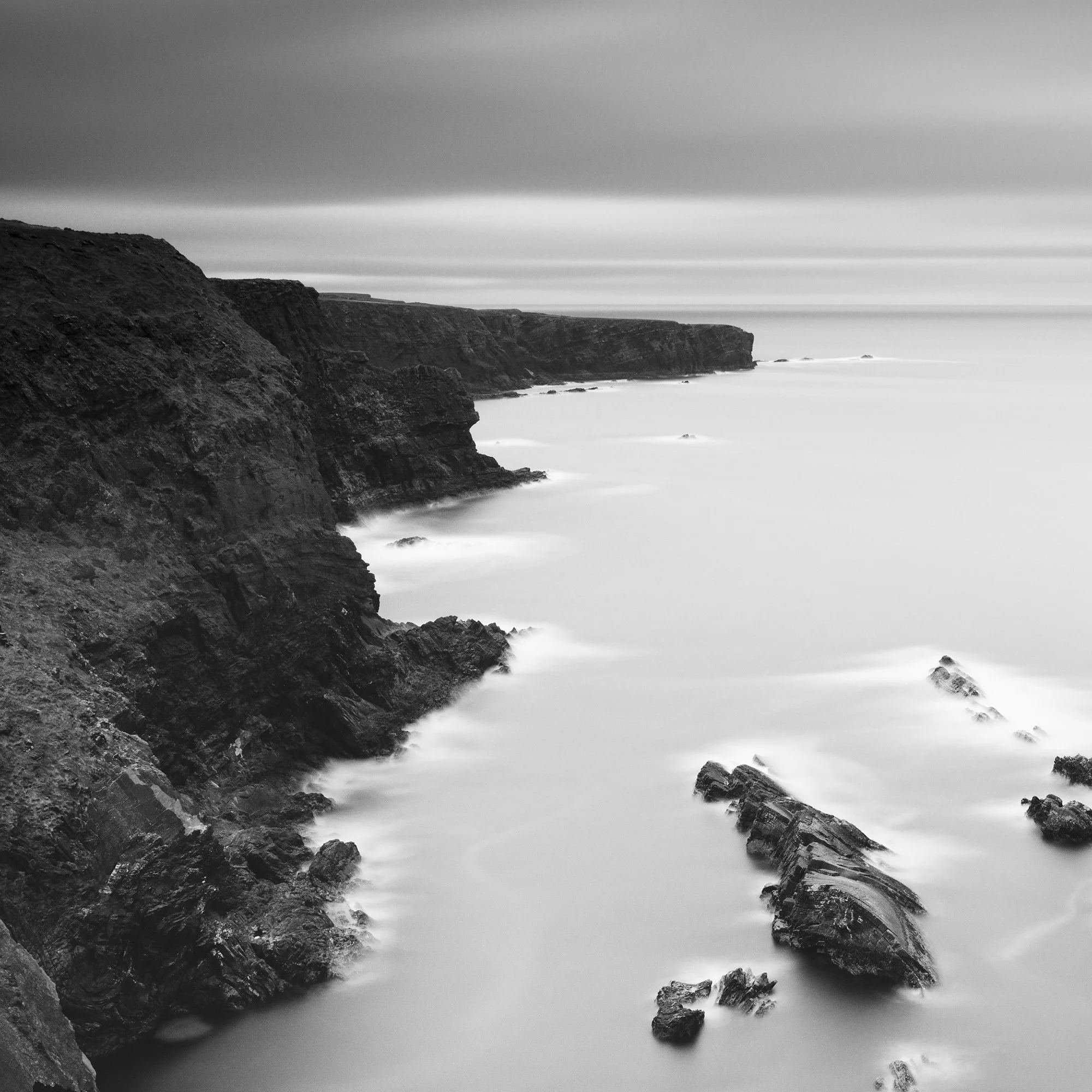 Gerald Berghammer - Black and white panorama seascape photography. Rocky coastline with cliffs and a tall rock formation in the water, under a cloudy sky. Print detail 1