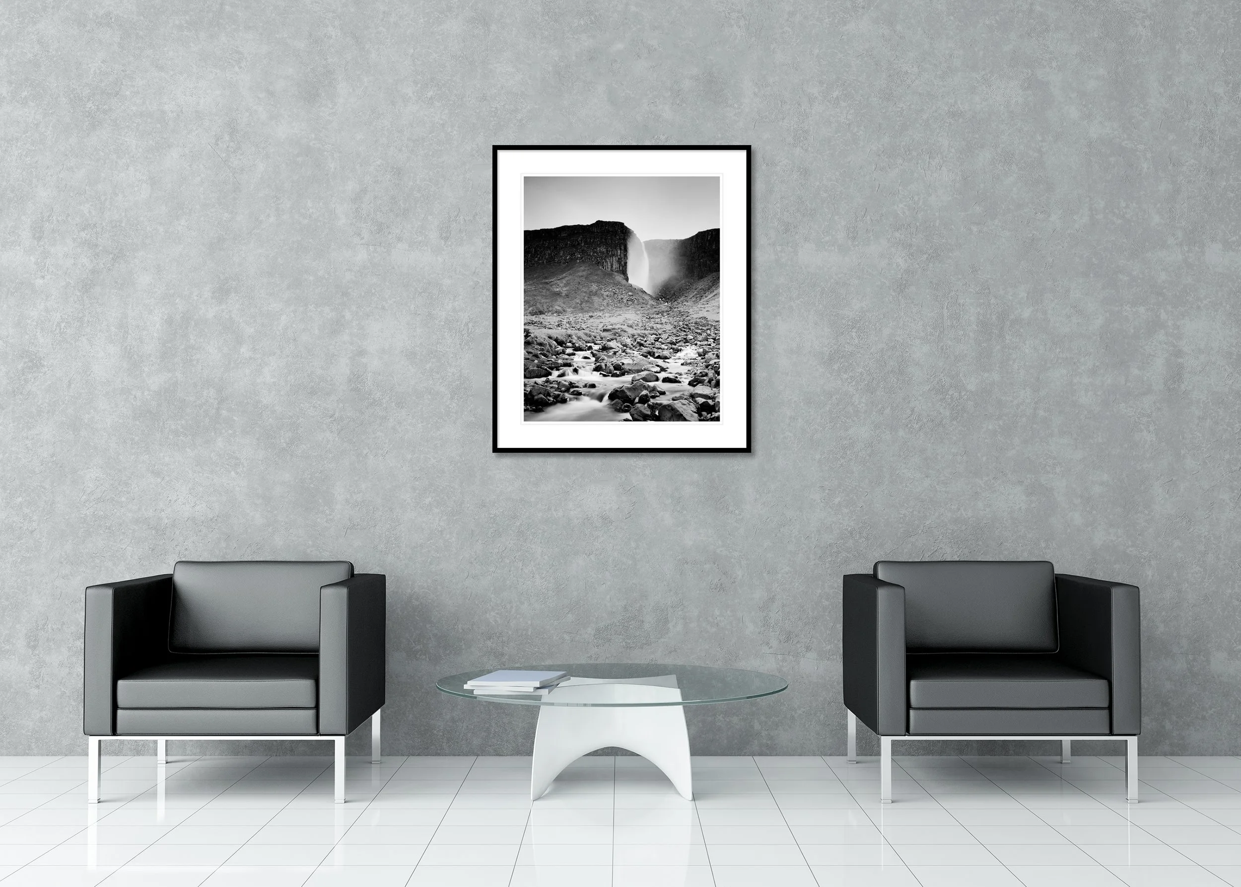 Monochrome landscape of an Icelandic waterfall between cliffs with misty spray and a rugged boulder-strewn riverbed, archival pigment print in a classic frame with mount.