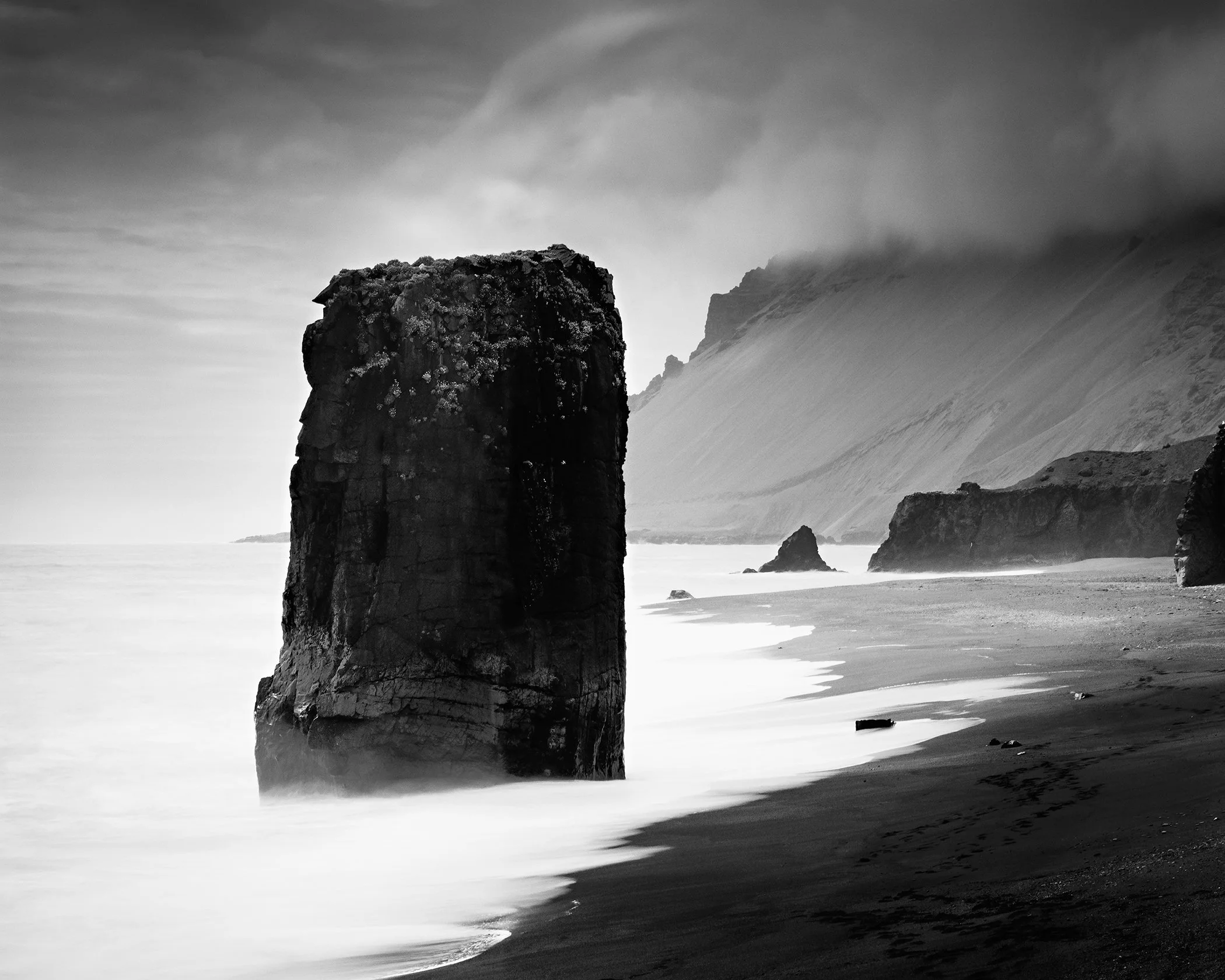 Black-and-white long-exposure photo of rock monolith in sea, foggy Iceland coasts with dark sand and misty water.