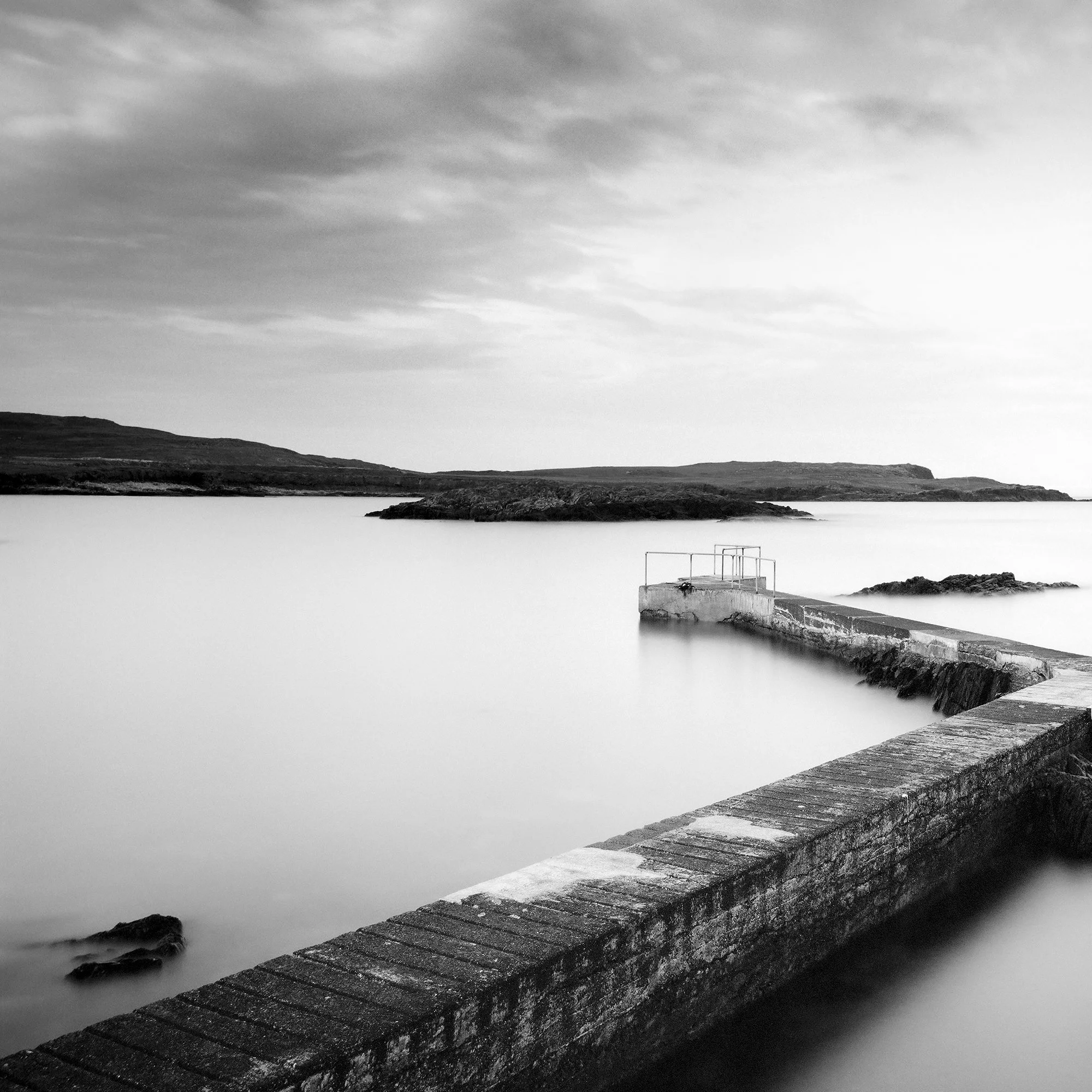© 2014 Gerald Berghammer - Black and white seascape photography. A Concrete pier extends into calm seawater with rocky edges, under a cloudy sky. Print detail 2