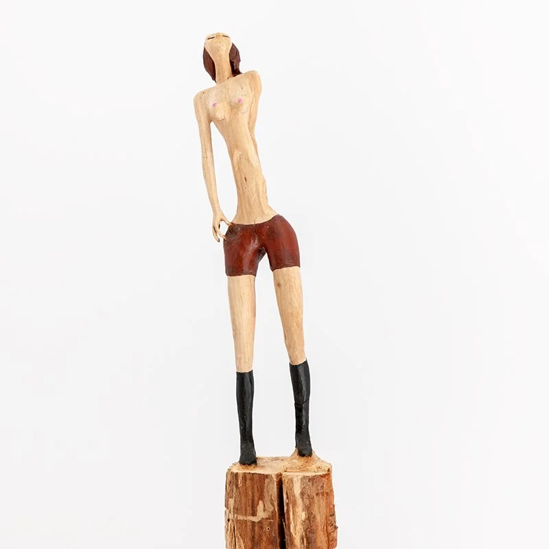 Wooden sculpture of a slender woman with short brown hair, wearing red shorts and black boots, standing on a wooden base.