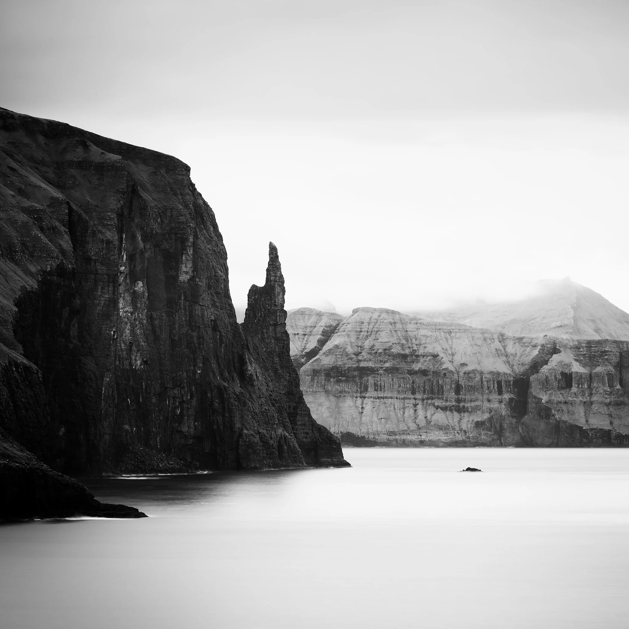 Trøllkonufingur Monolith Study 2, black-and-white long-exposure photograph of dramatic sea cliffs, a slender rock formation and misty mountains in the Faroe Islands by Gerald Berghammer.