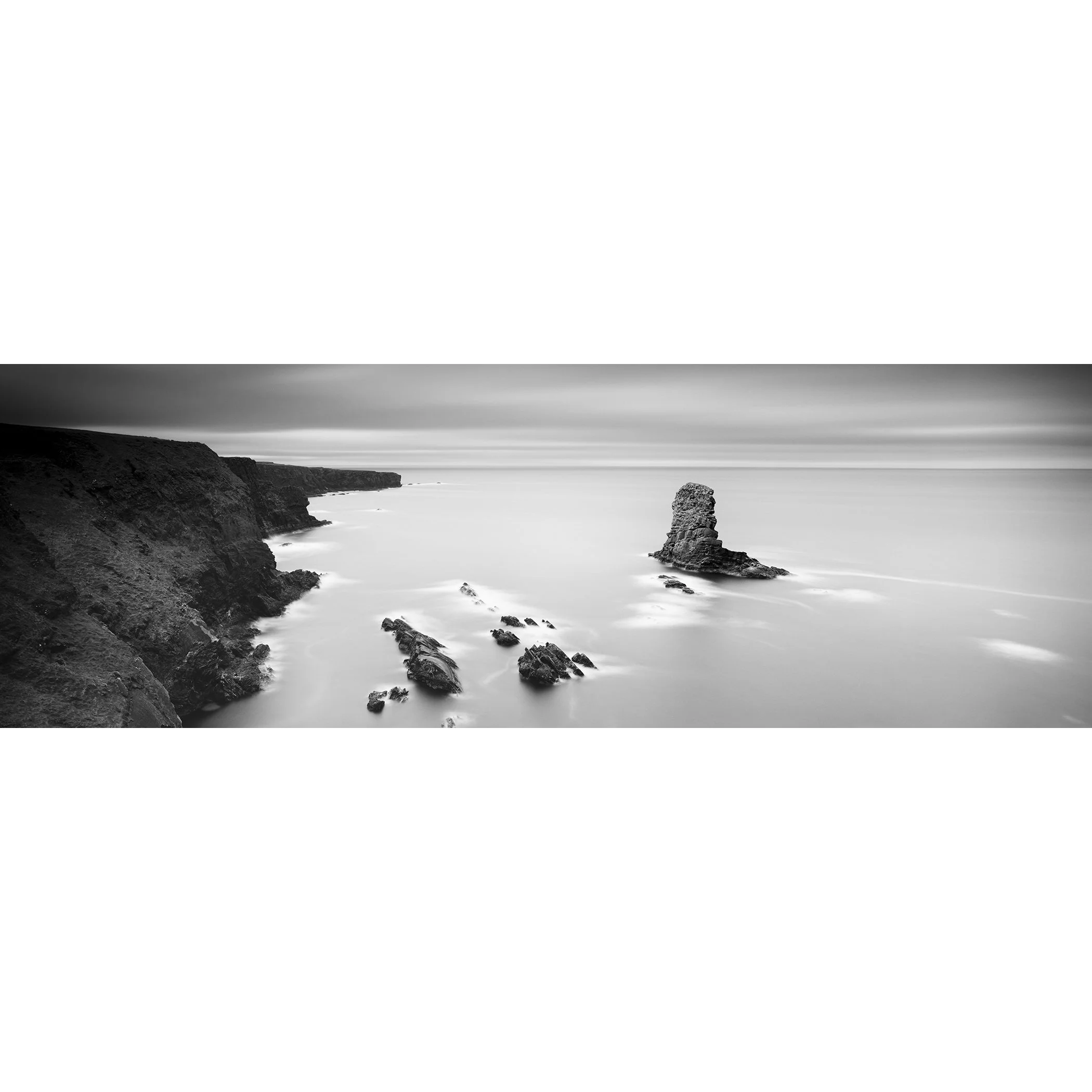 Gerald Berghammer - Black and white panorama seascape photography. Rocky coastline with cliffs and a tall rock formation in the water, under a cloudy sky.