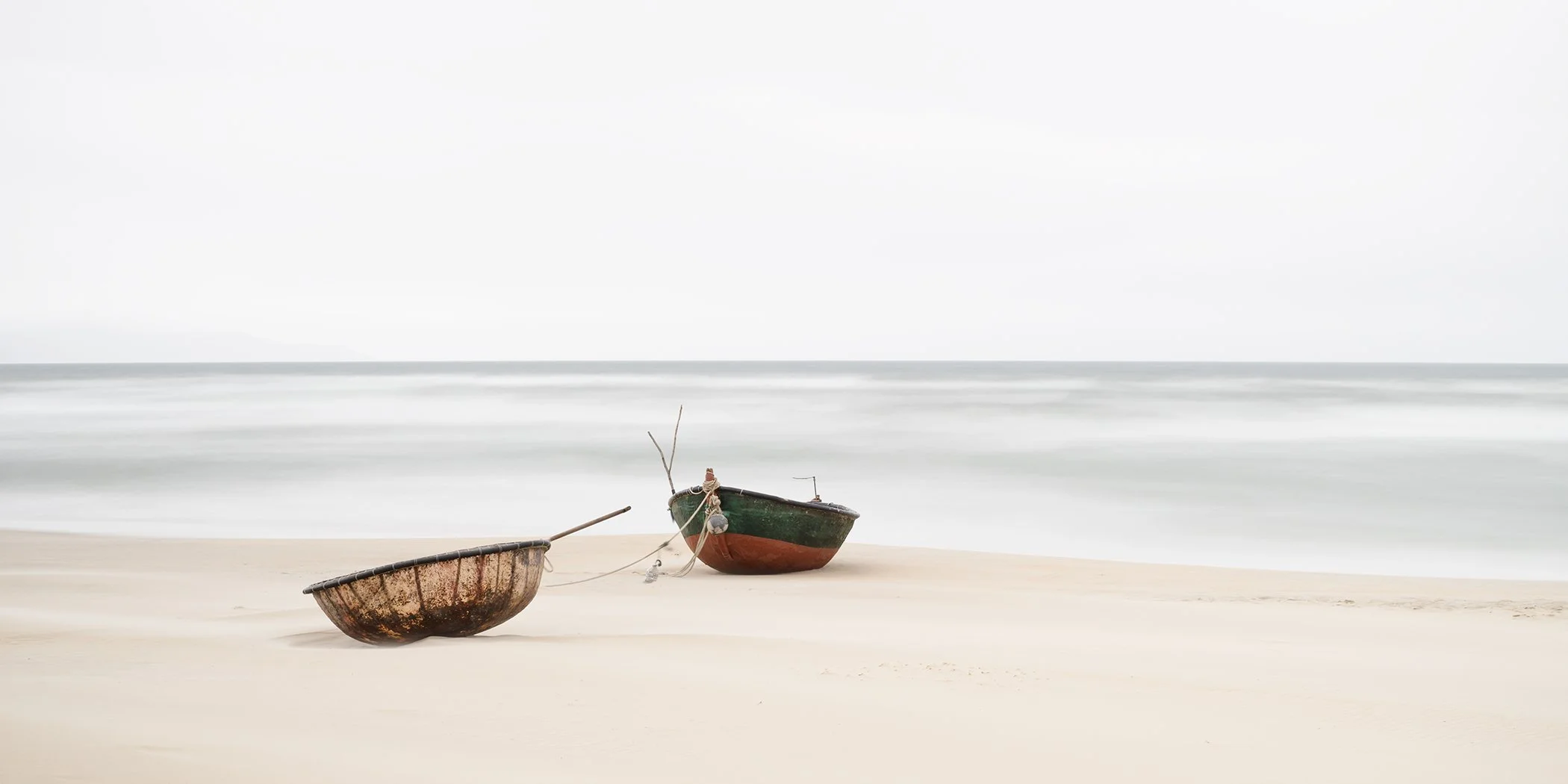 Two small fishing boats on a quiet sandy beach, with a calm sea and pale overcast sky in a minimalist coastal scene