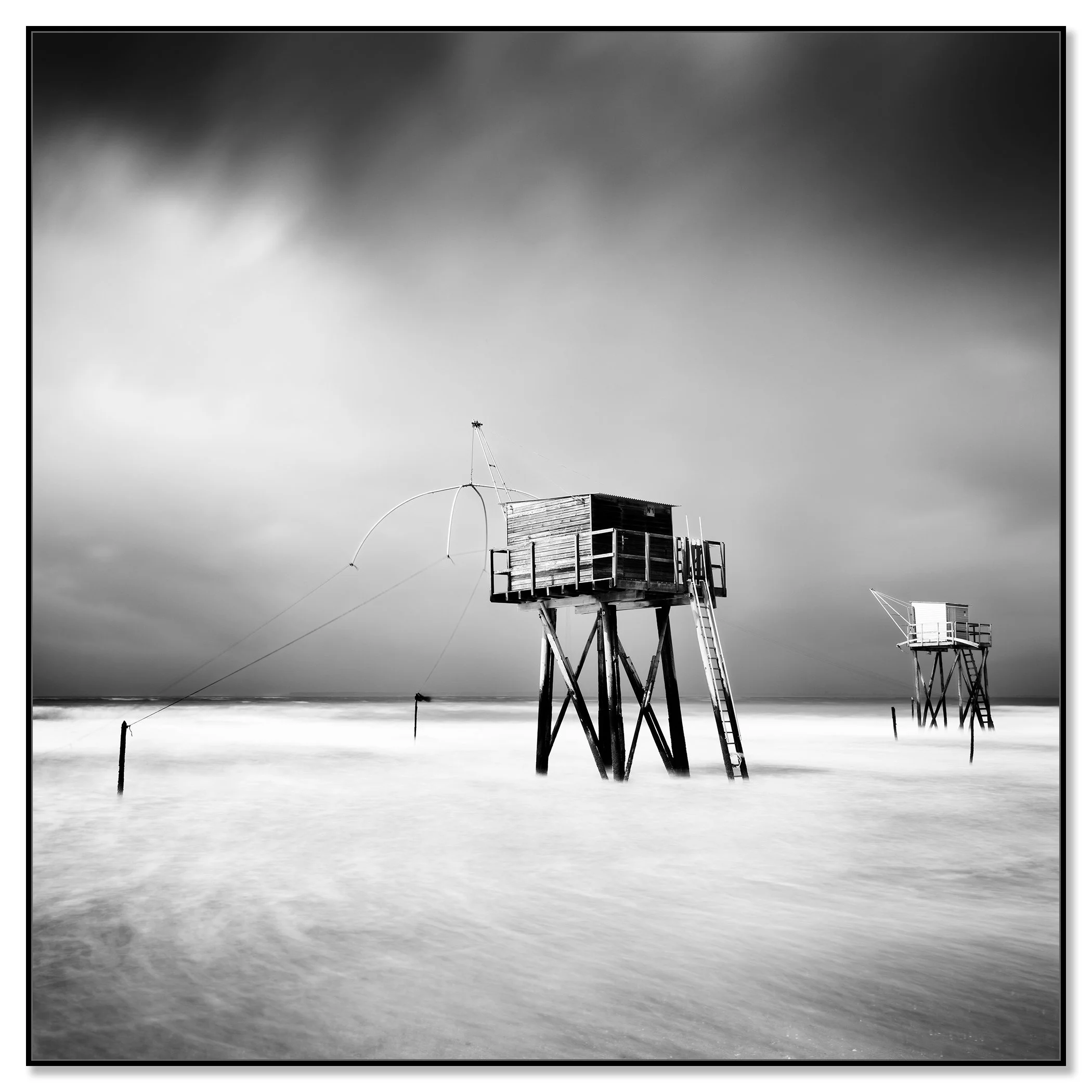 Black and white long-exposure photo of wooden fishing huts on stilts, reached by ladders, over calm sea water under a dramatic cloudy sky – framed ArtBox black