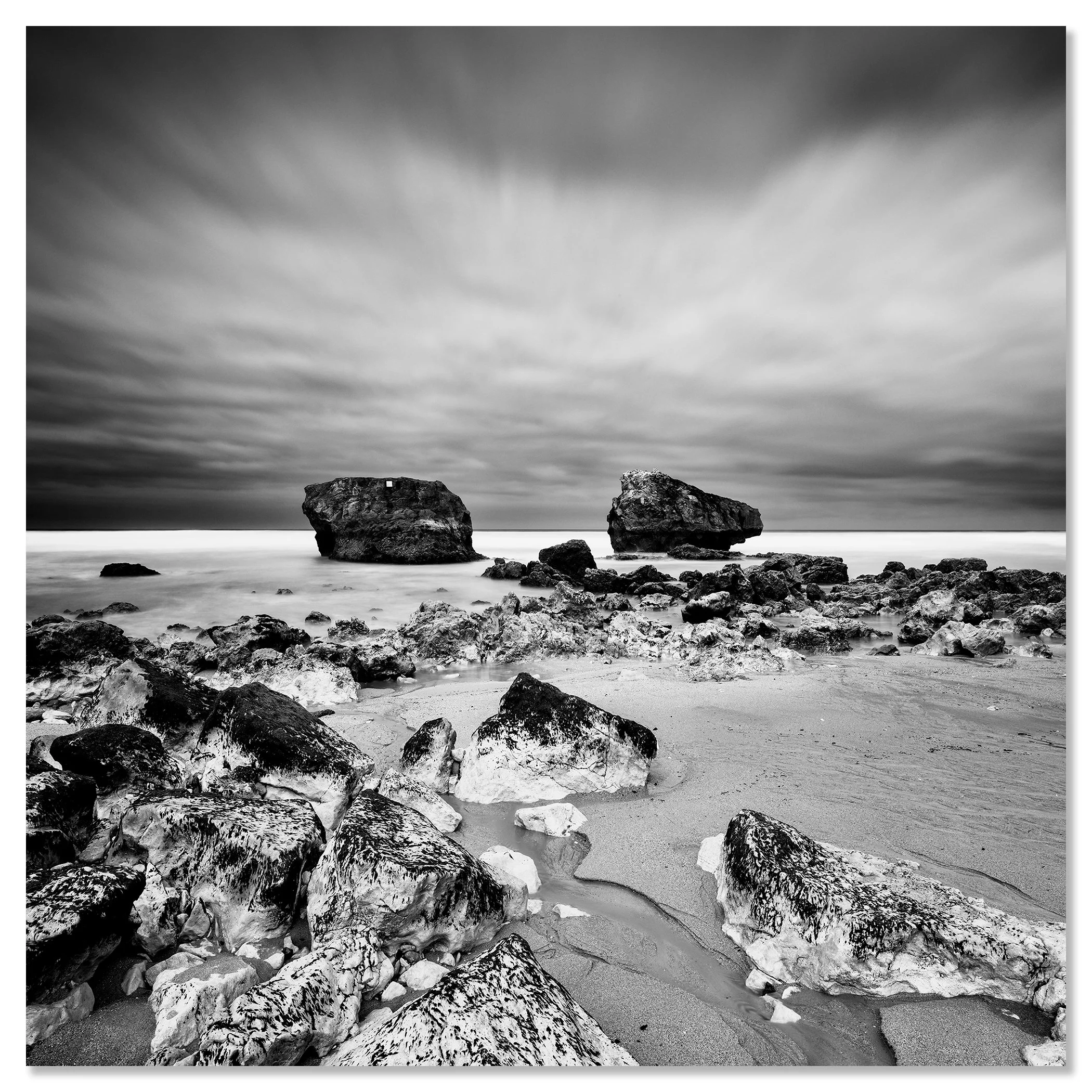 Black-and-white rocky beach with boulders offshore under dramatic, streaked clouds – dibond frameless