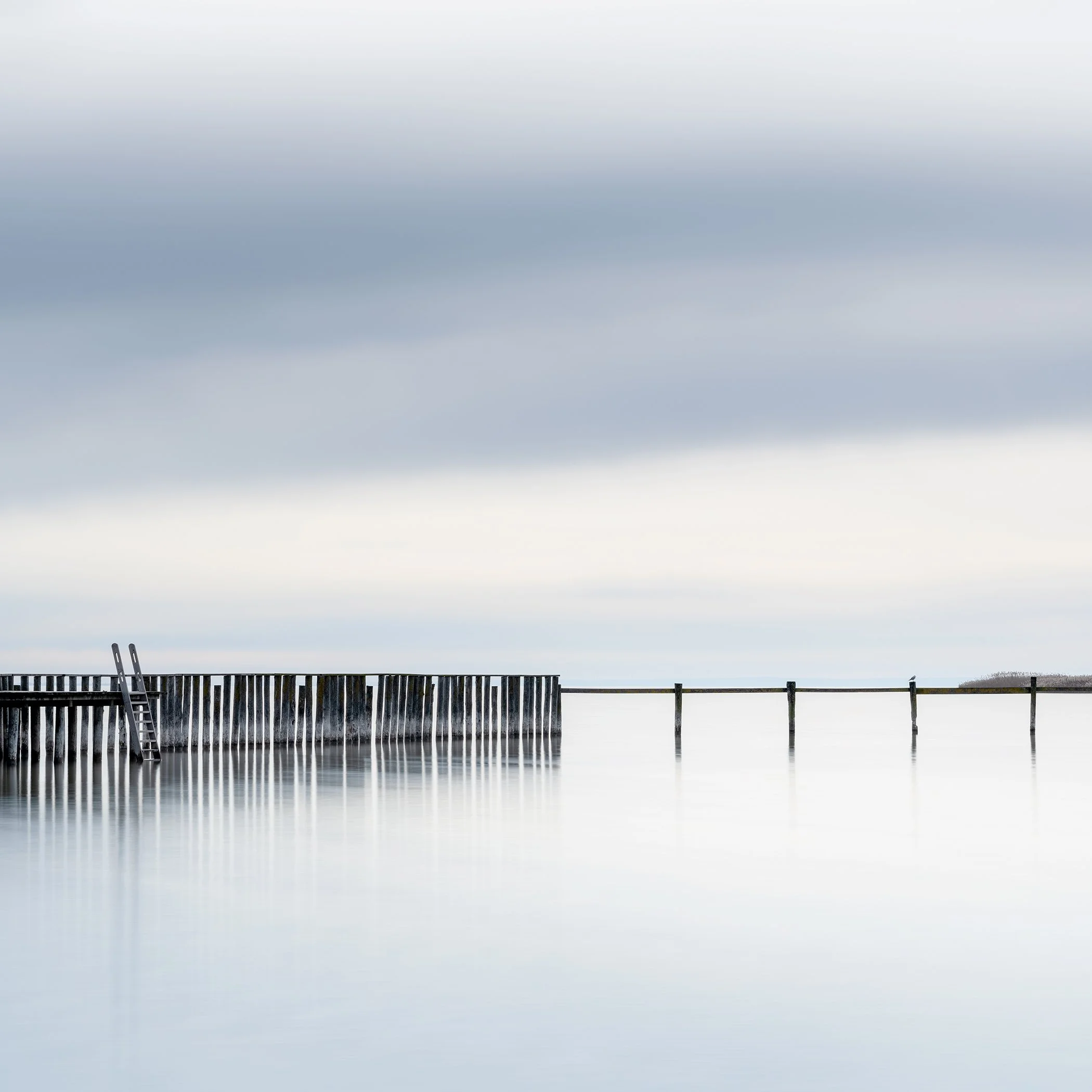 Minimalist colour photograph of a quiet swimming area with a wooden barrier and bathing ladder in Austria. Photo by Gerald Berghammer.