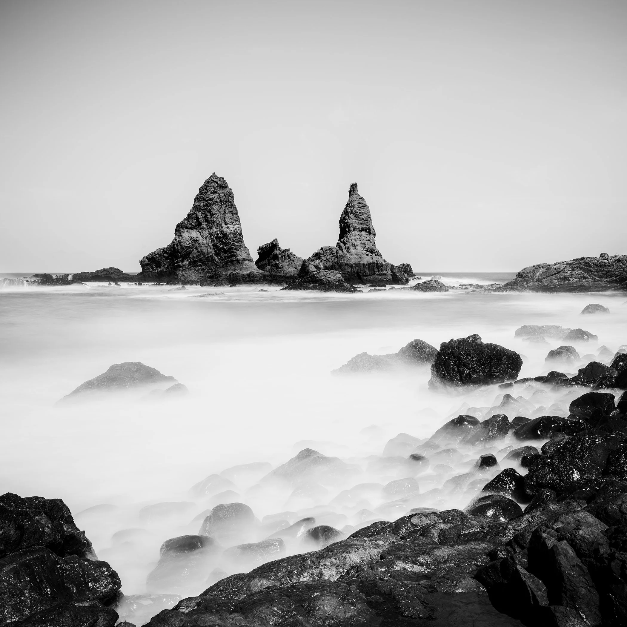 Long-exposure black-and-white coastal photograph showing sea stacks rising from calm mist and dark shoreline rocks.