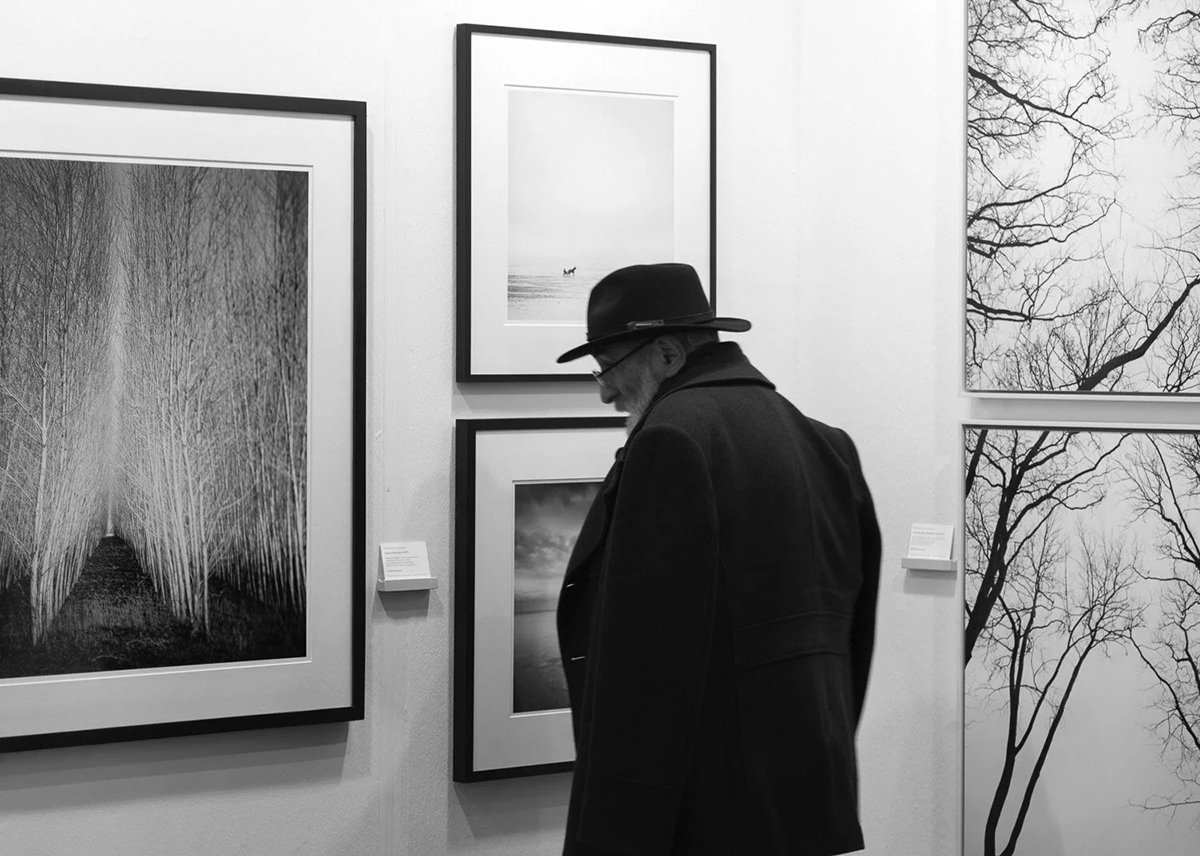 Visitor wearing a dark hat beside black-and-white forest and tree photographs by Gerald Berghammer at art KARLSRUHE 2018.