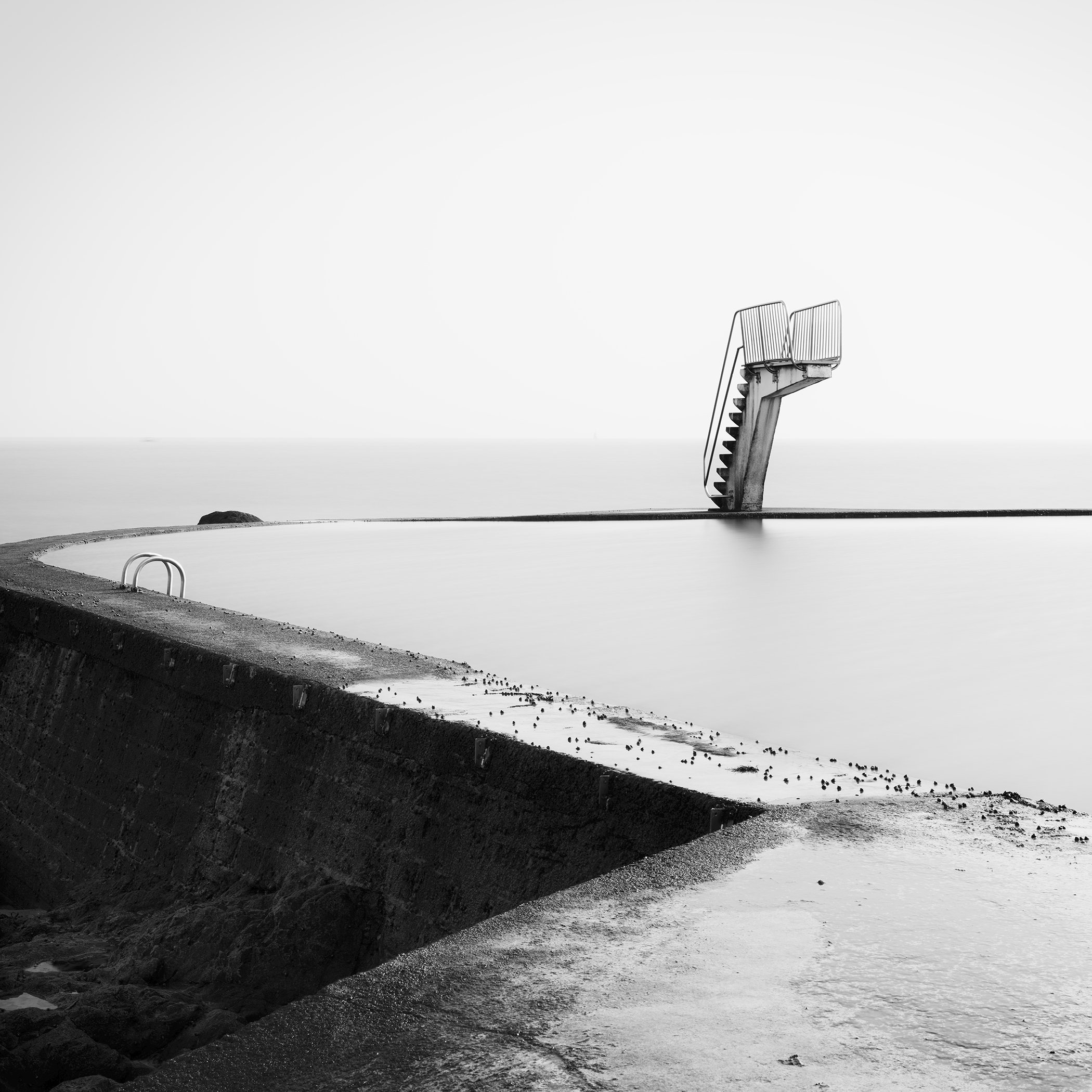 Minimalist monochrome image of a seawater pool with a diving platform and smooth, still water.