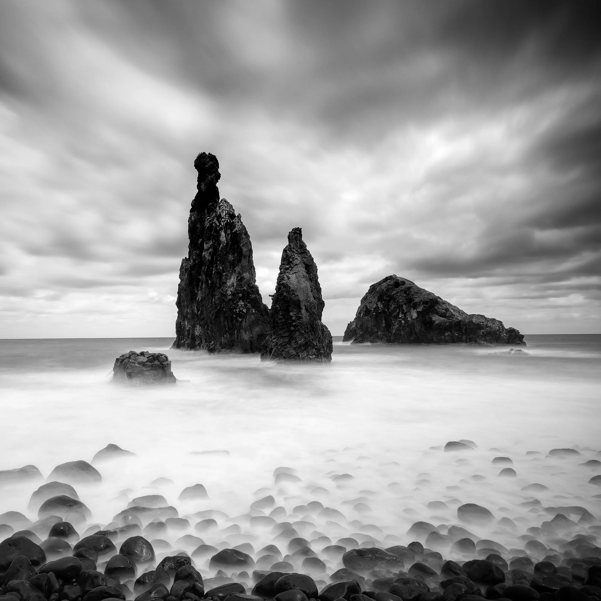 © 2021 Gerald Berghammer - Black and white seascape photography. Rocky seashore with three prominent sea stacks rising from the water, cloudy sky overhead. Print detail 2