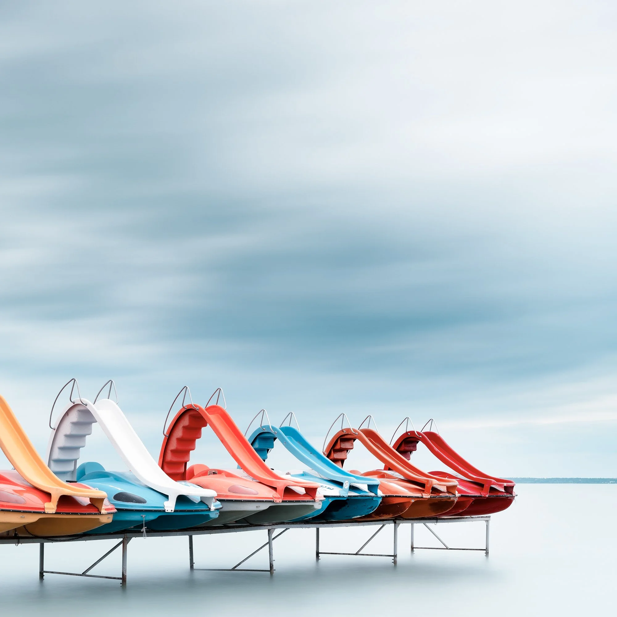Gerald Berghammer - Color waterscape photography. A row of colorful plastic pedal boats shaped like race cars on a dock over water, under a cloudy sky. Print detail 2