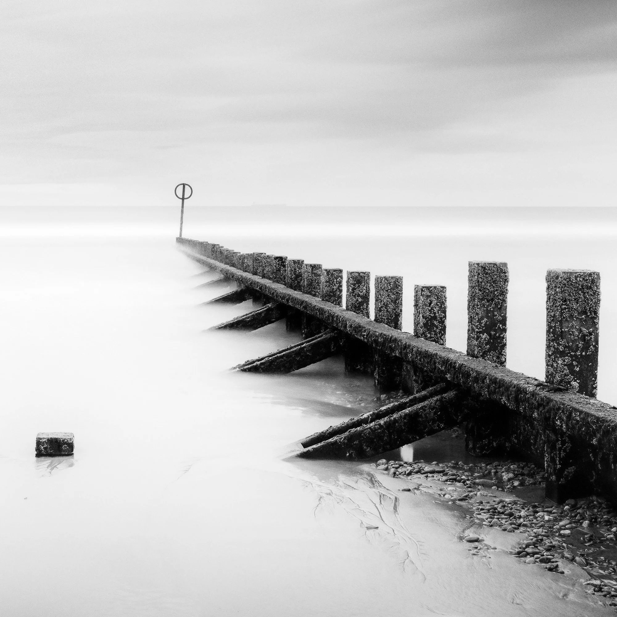 © 2012 Gerald Berghammer - Black and white landscape photography. A wooden wave breaker extending into a calm body of water, with a circular marker at the end. Print detail 3