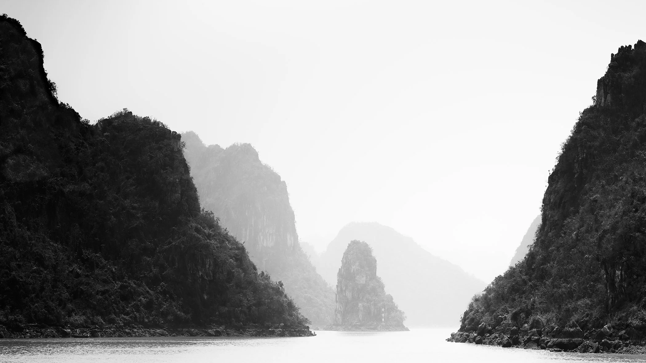 Black-and-white photo of a calm sea passage between steep, forested limestone cliffs, with misty mountains in the background and still water in the foreground.