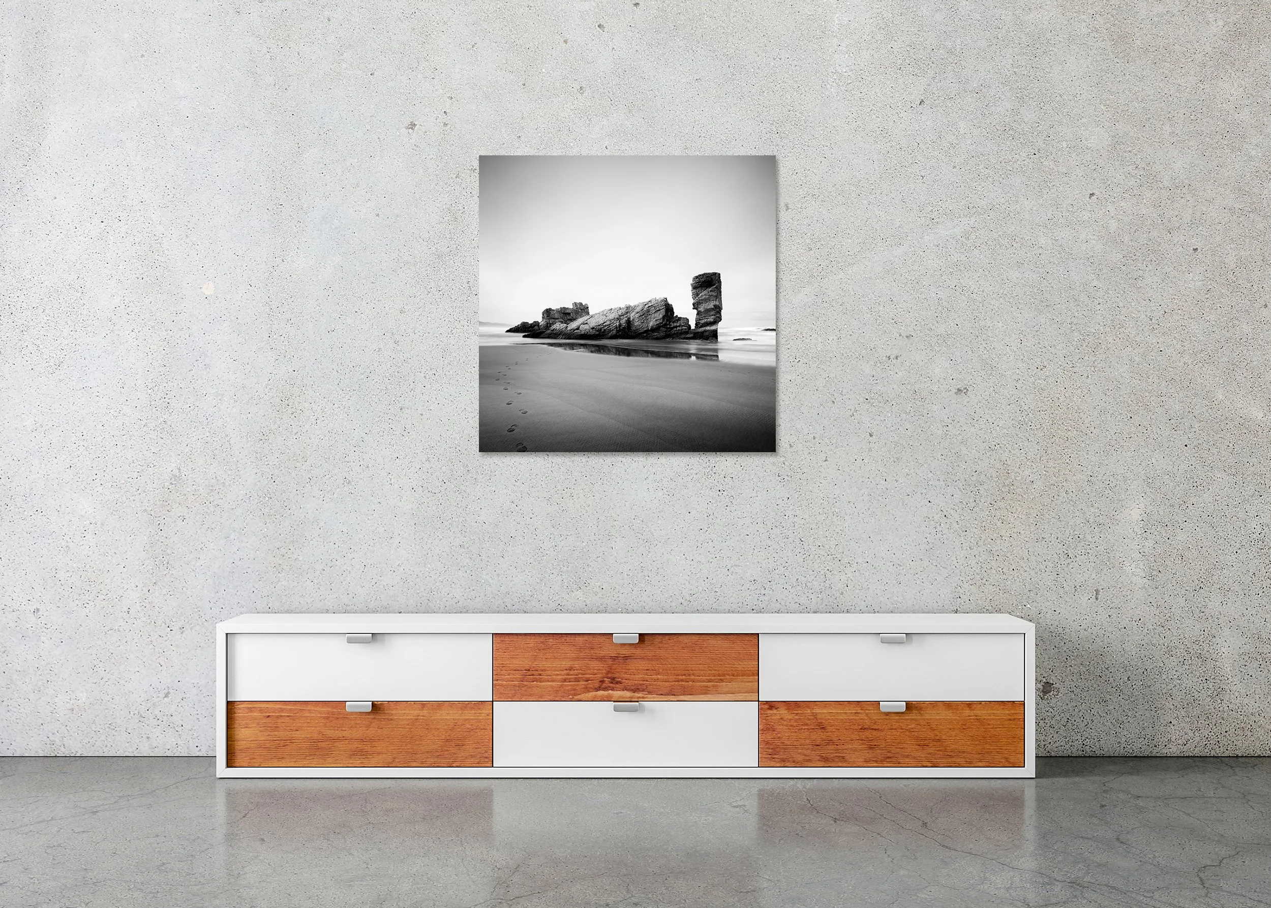 Black-and-white seaside view with damp sand, faint footprints and a towering shore rock reflected in calm shallow water, photo print mounted to frameless Dibond