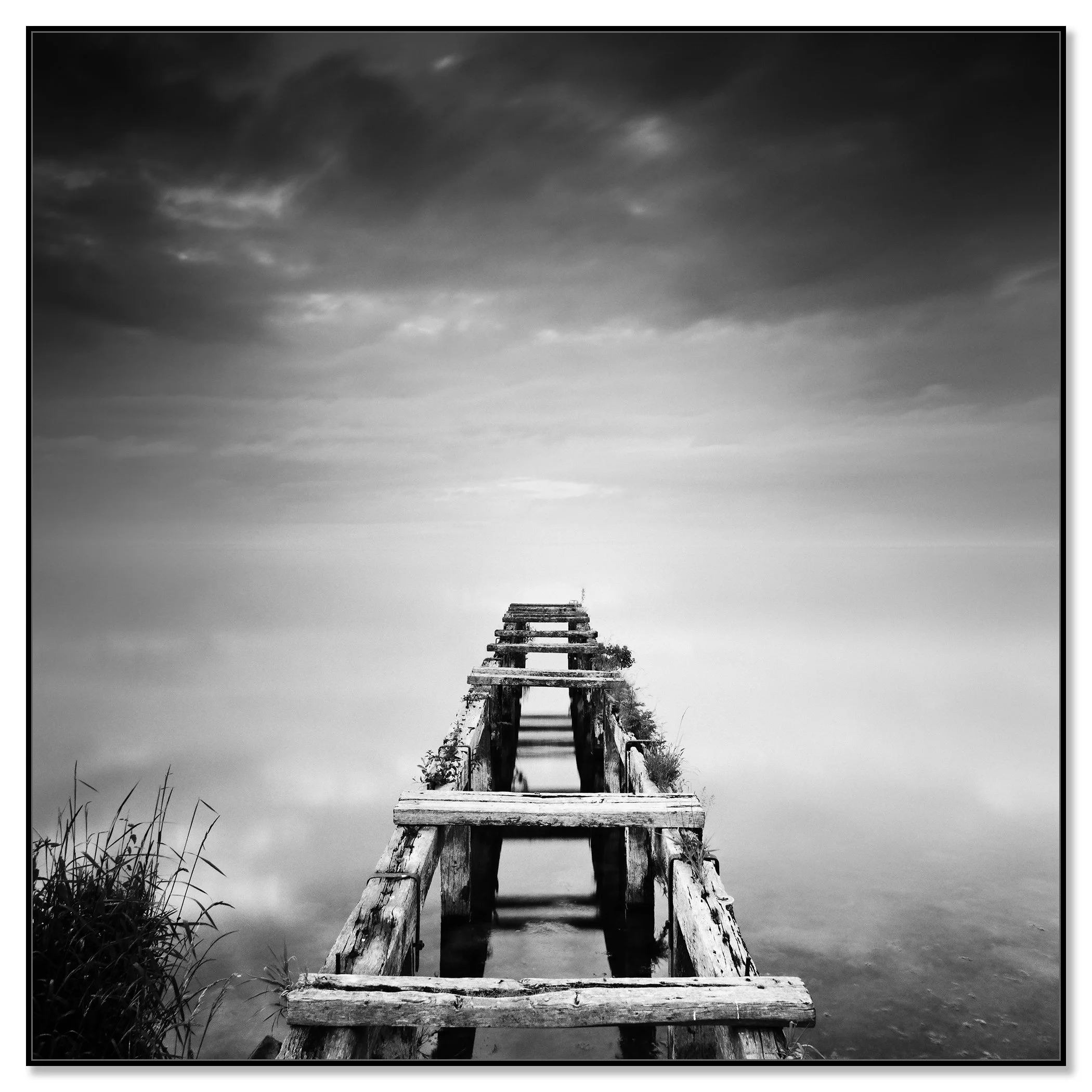 Gerald Berghammer - Black and white waterscape photography. An old abandoned wooden dock extending into a calm body of water under a cloudy sky. Chromaluxe framed black