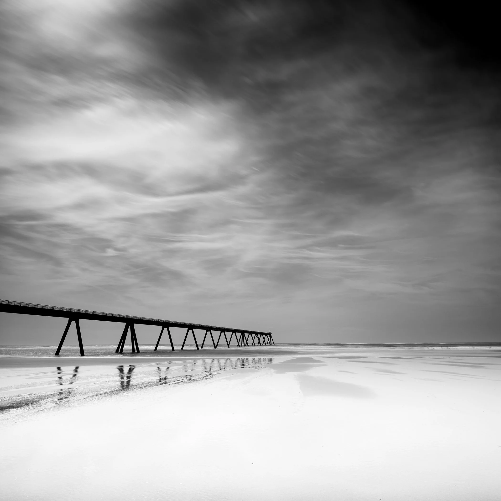Black and white photograph of a long wooden pier on a calm beach beneath a dramatic cloudy sky