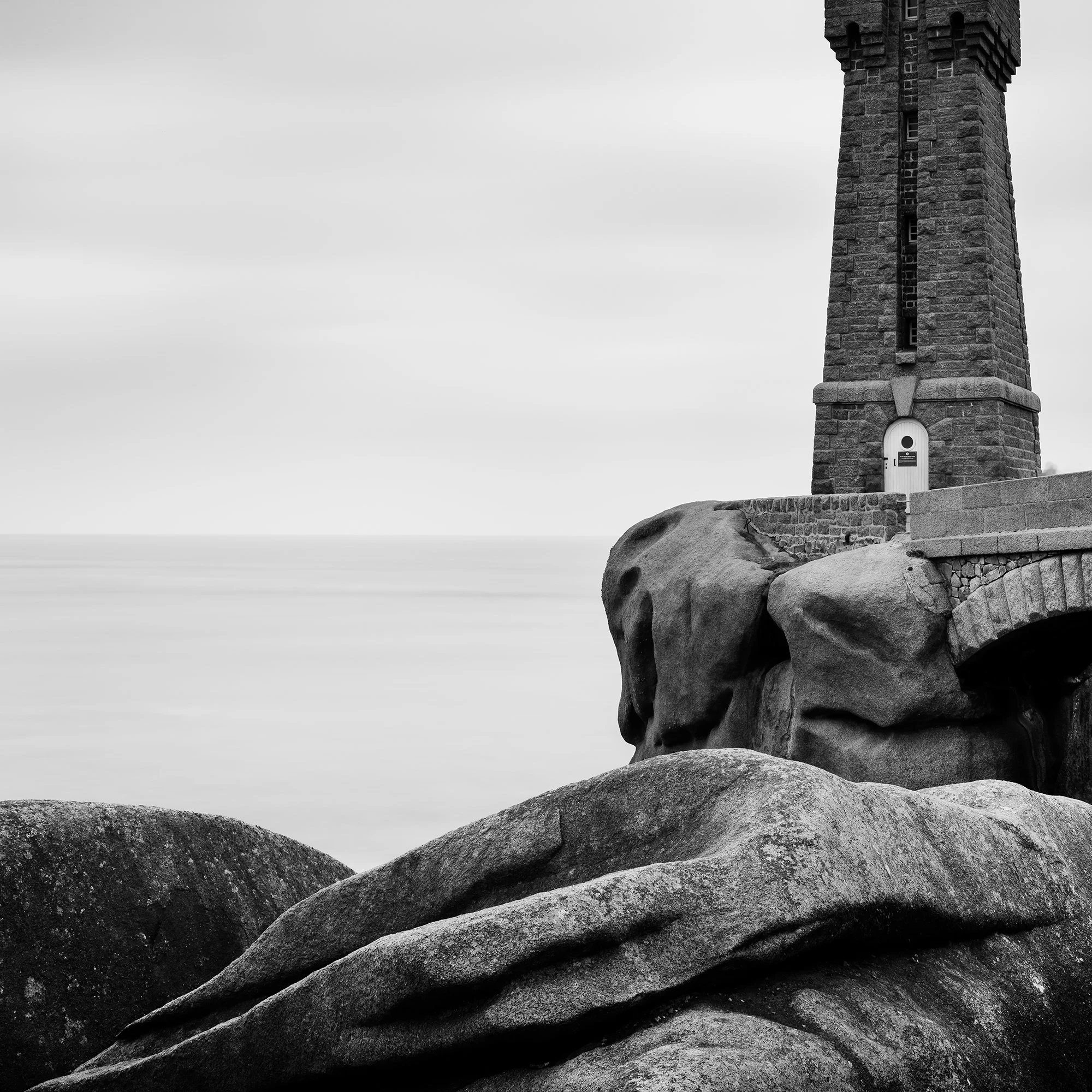 Gerald Berghammer - Black and white landscape photograph of a tall lighthouse on a rocky coast, with a small bridge and large rocks in foreground. Print detail 1