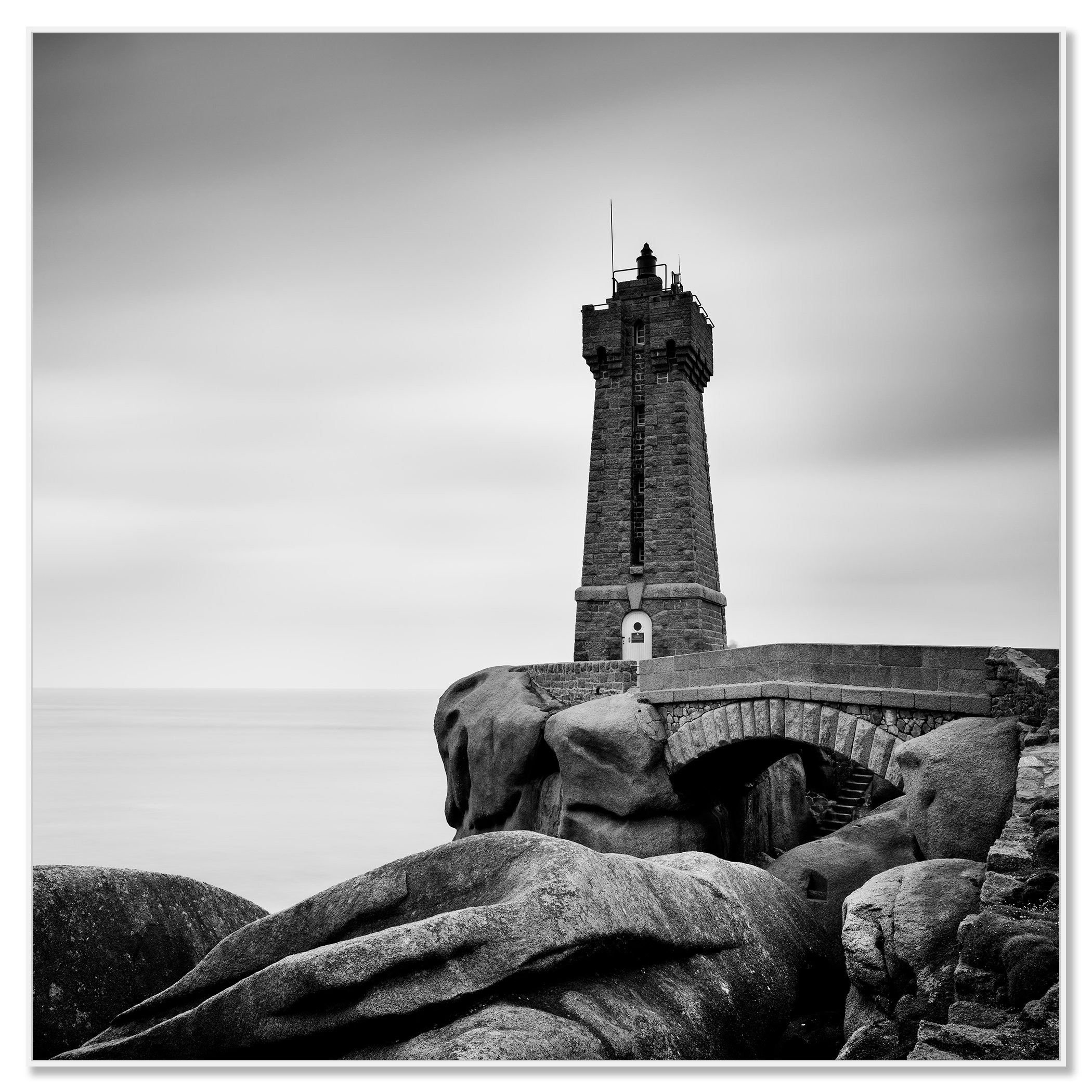 Gerald Berghammer - Black and white landscape photograph of a tall lighthouse on a rocky coast, with a small bridge and large rocks in foreground. Chromaluxe framed white
