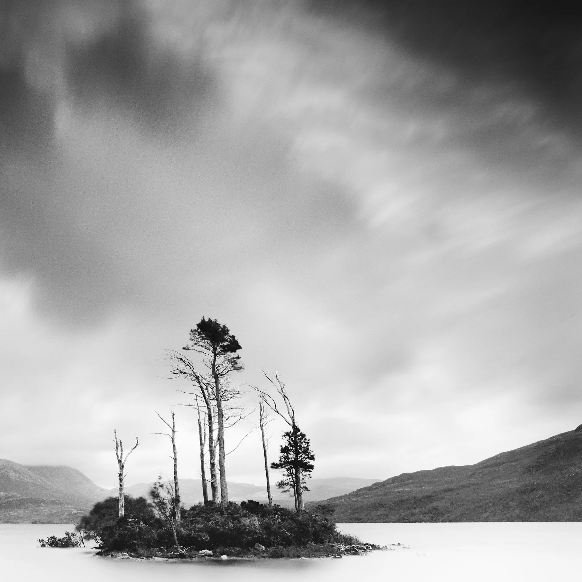 © 2012 Gerald Berghammer - Black and white waterscape photography. Small island with tall, thin trees surrounded by water and distant mountains under a cloudy sky. Print detail 2