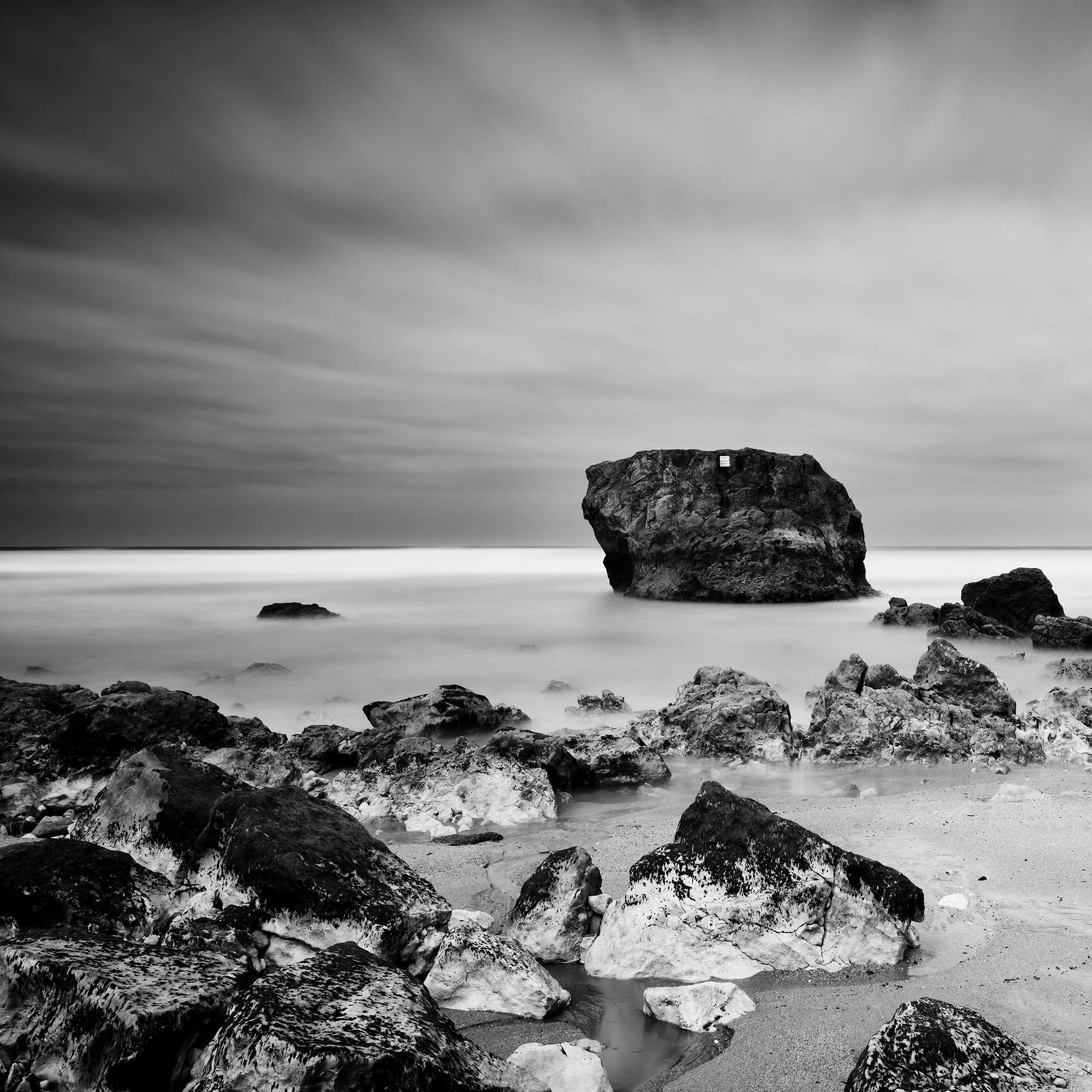 Gerald Berghammer - Black and white landscape photo. Rocky beach with two large rocks protruding from the water, cloudy sky, and smooth ocean surface. Print detail 1