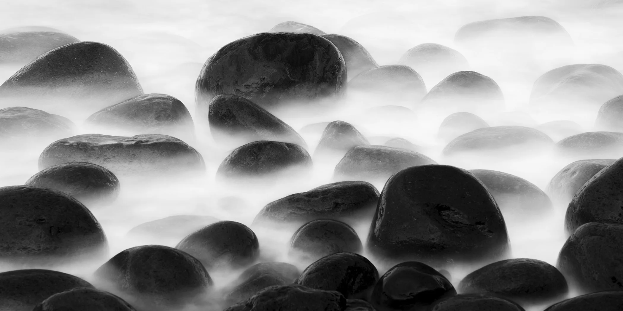 Black and white fine art photograph by Gerald Berghammer showing smooth coastal rocks surrounded by misty long-exposure seawater.