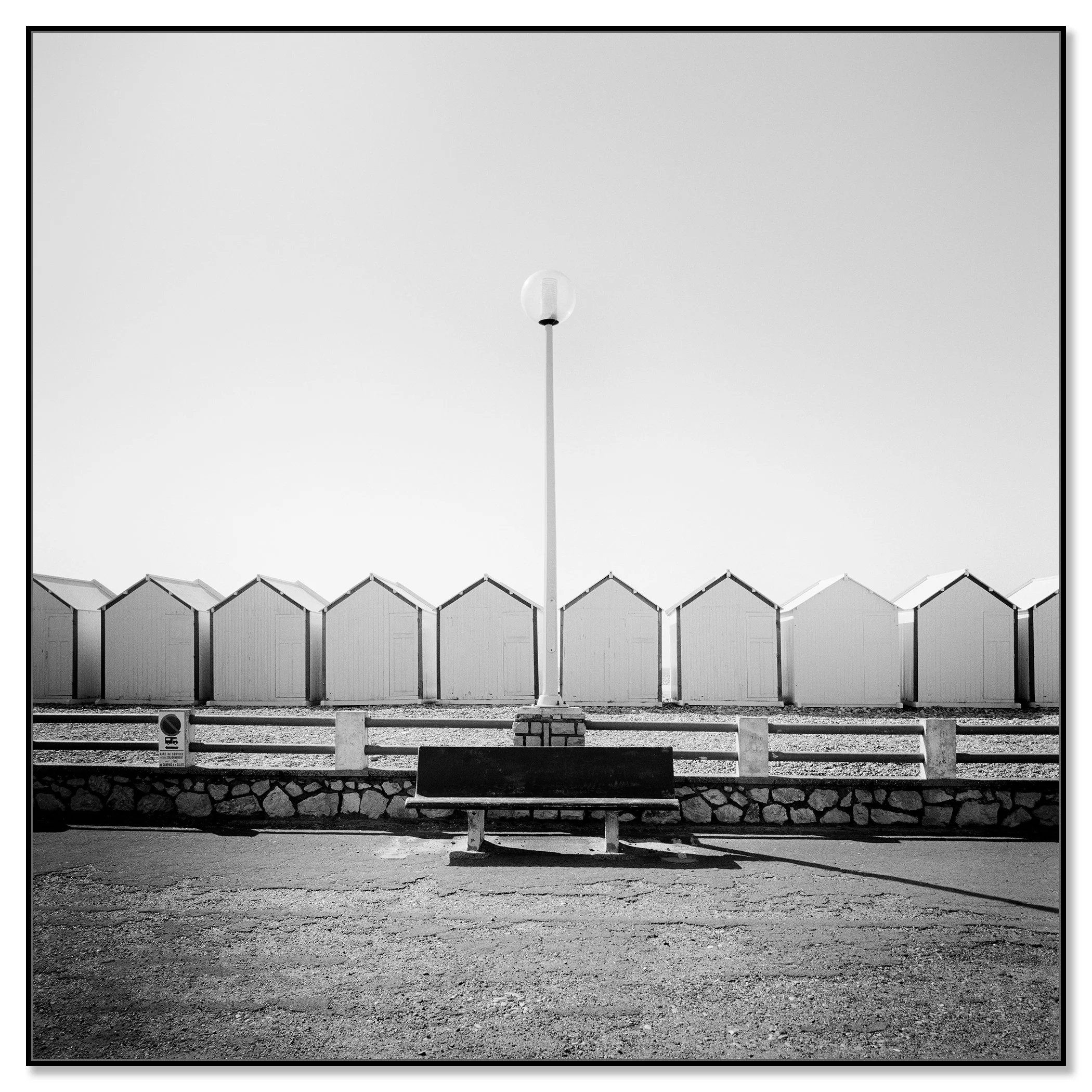 Empty bench on promenade facing beach huts, centred streetlamp; minimalist black-and-white coastal photo – framed ArtBox black