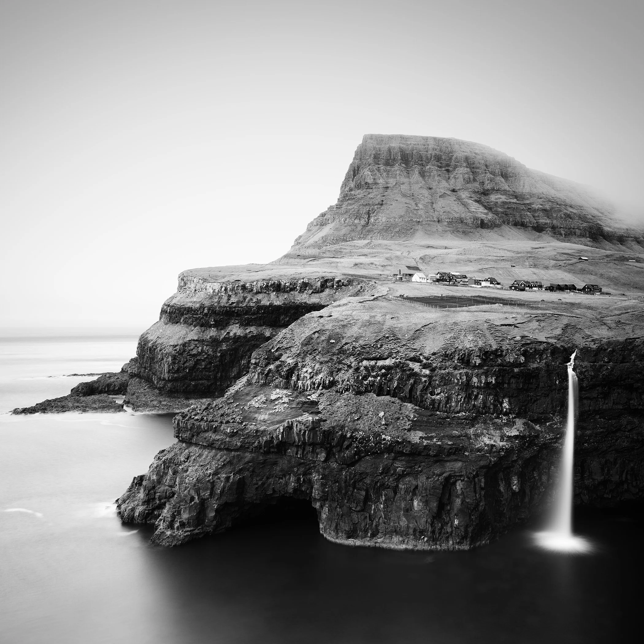 Black-and-white photo of a layered coastal cliff with a narrow waterfall flowing into the ocean, and a small settlement on the plateau above.